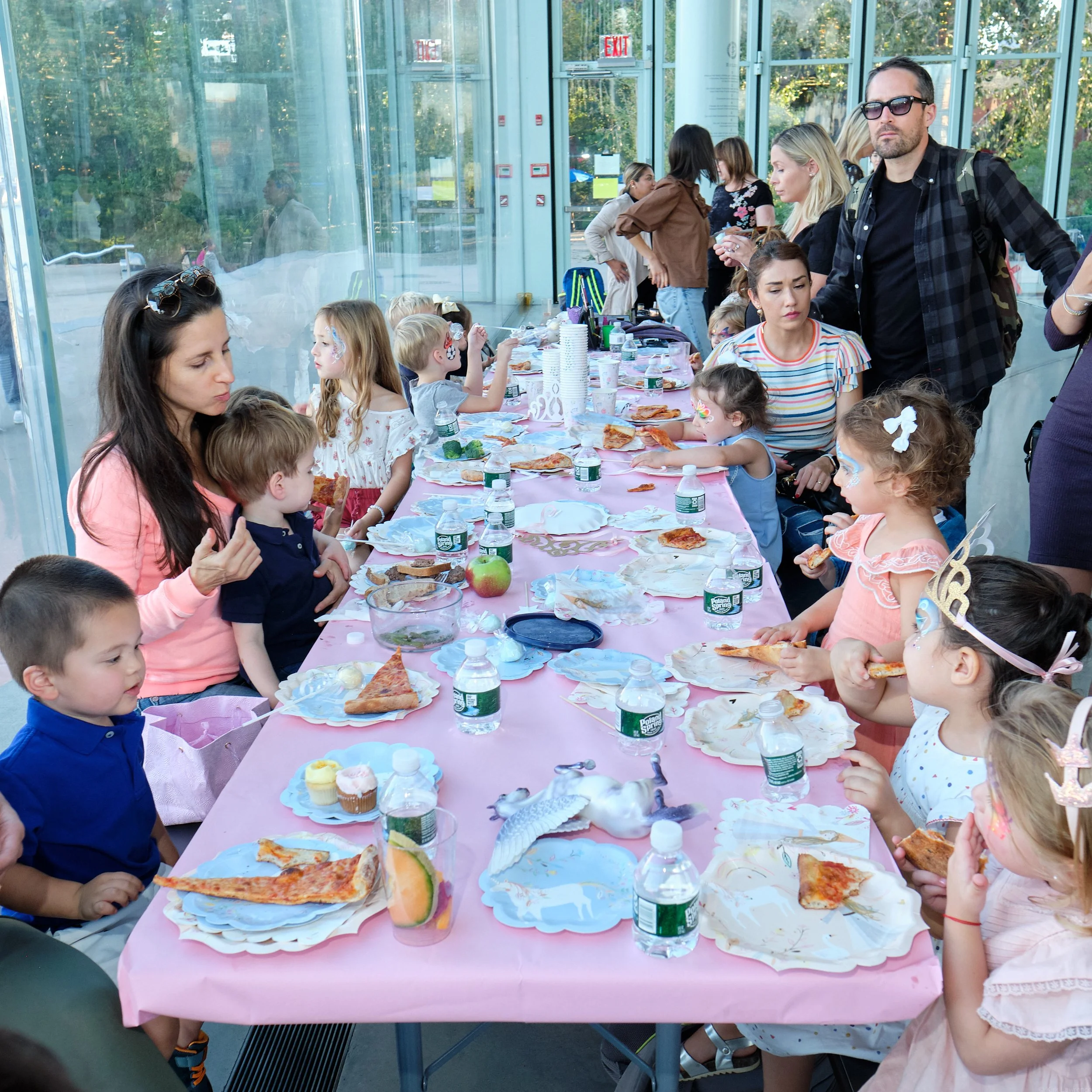 Children and adults sit at a long pink table inside a glass-walled building, eating pizza and snacks during a celebration or party.