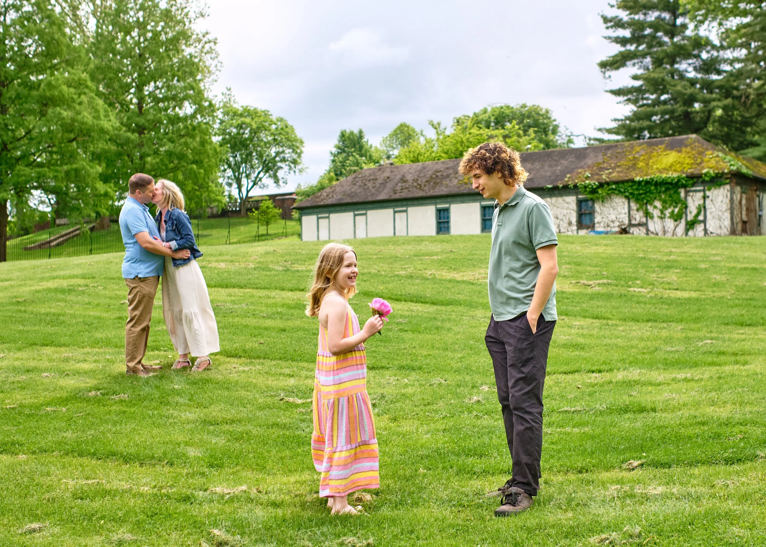 A young girl in a colorful striped dress holding pink flowers stands on a grassy field, smiling and talking with a teenage boy in a gray shirt and black pants. In the background, a couple is kissing, with green trees and a historic building with a mo