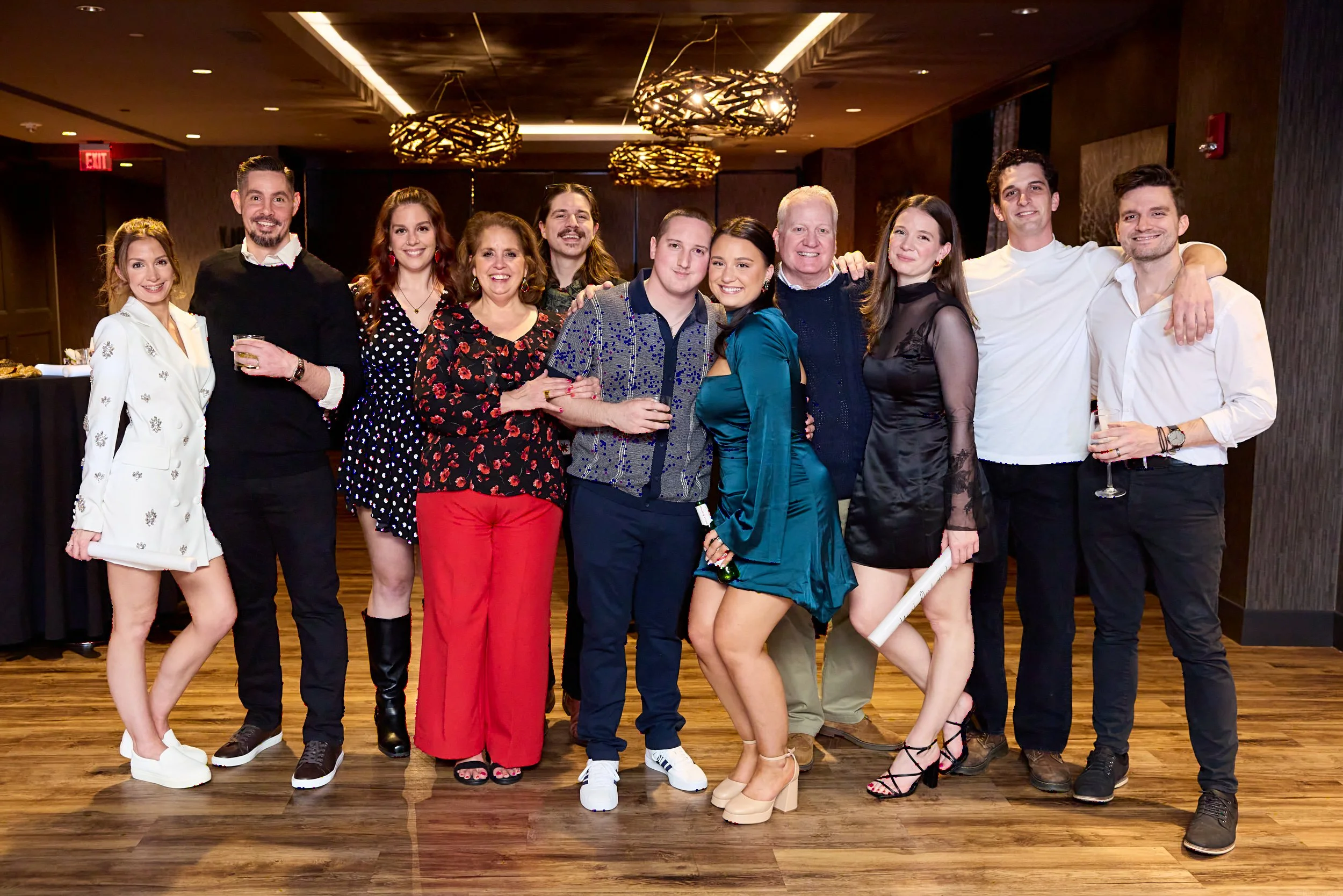 Group of twelve people at a social gathering, smiling and posing closely together indoors with warm lighting, hardwood floor, and contemporary decor.