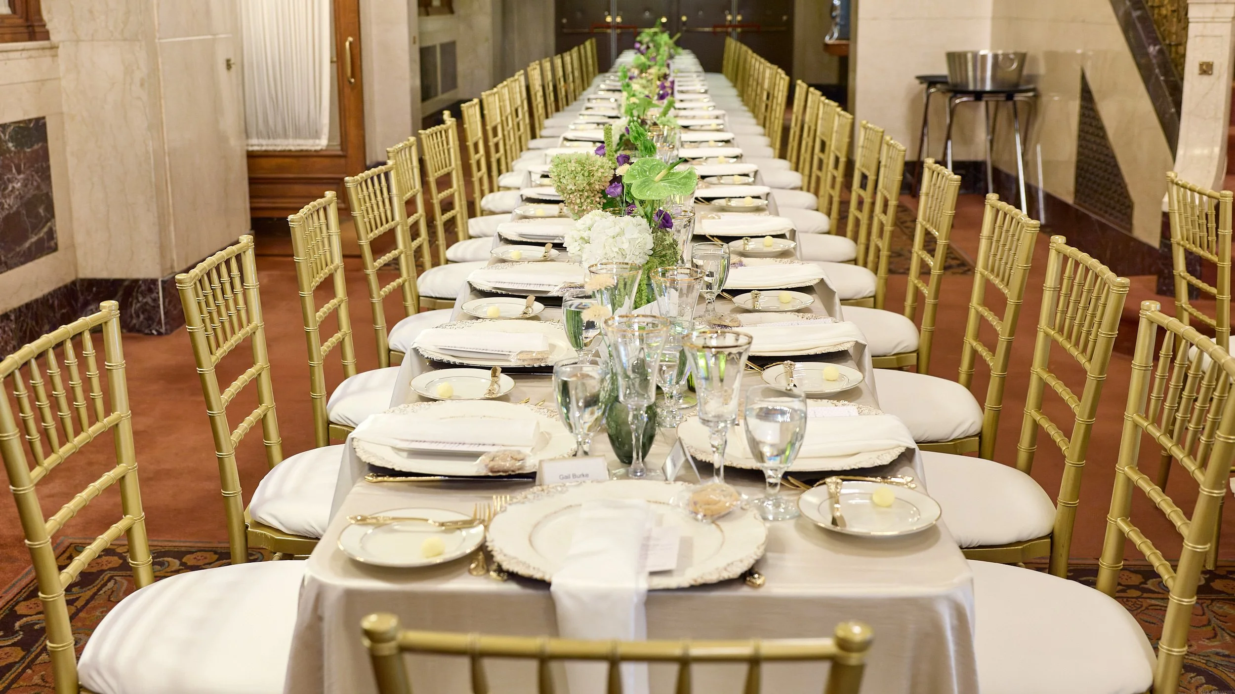 Long banquet table set for a formal event with white tablecloths, gold chairs, floral centerpieces, and place settings with plates, glasses, and utensils inside an elegant room.