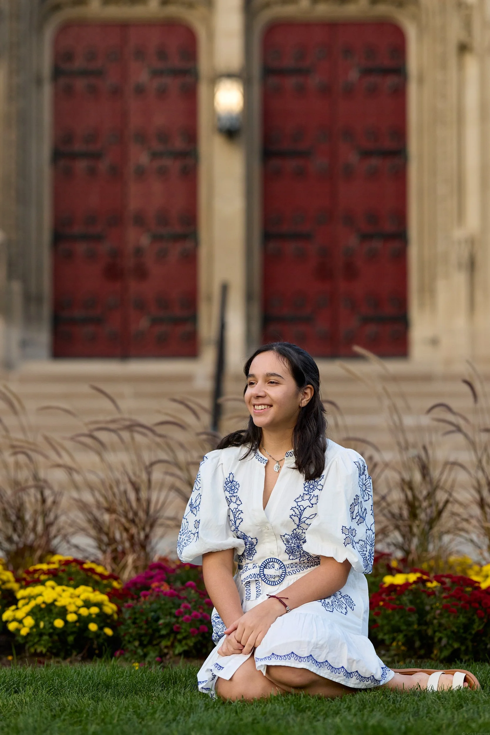 A smiling young woman kneeling on grass with her hands resting on her lap, wearing a white dress with blue embroidery, in front of a garden with colorful flowers and tall wild grasses, and an ornate building with large red doors and beige stone walls