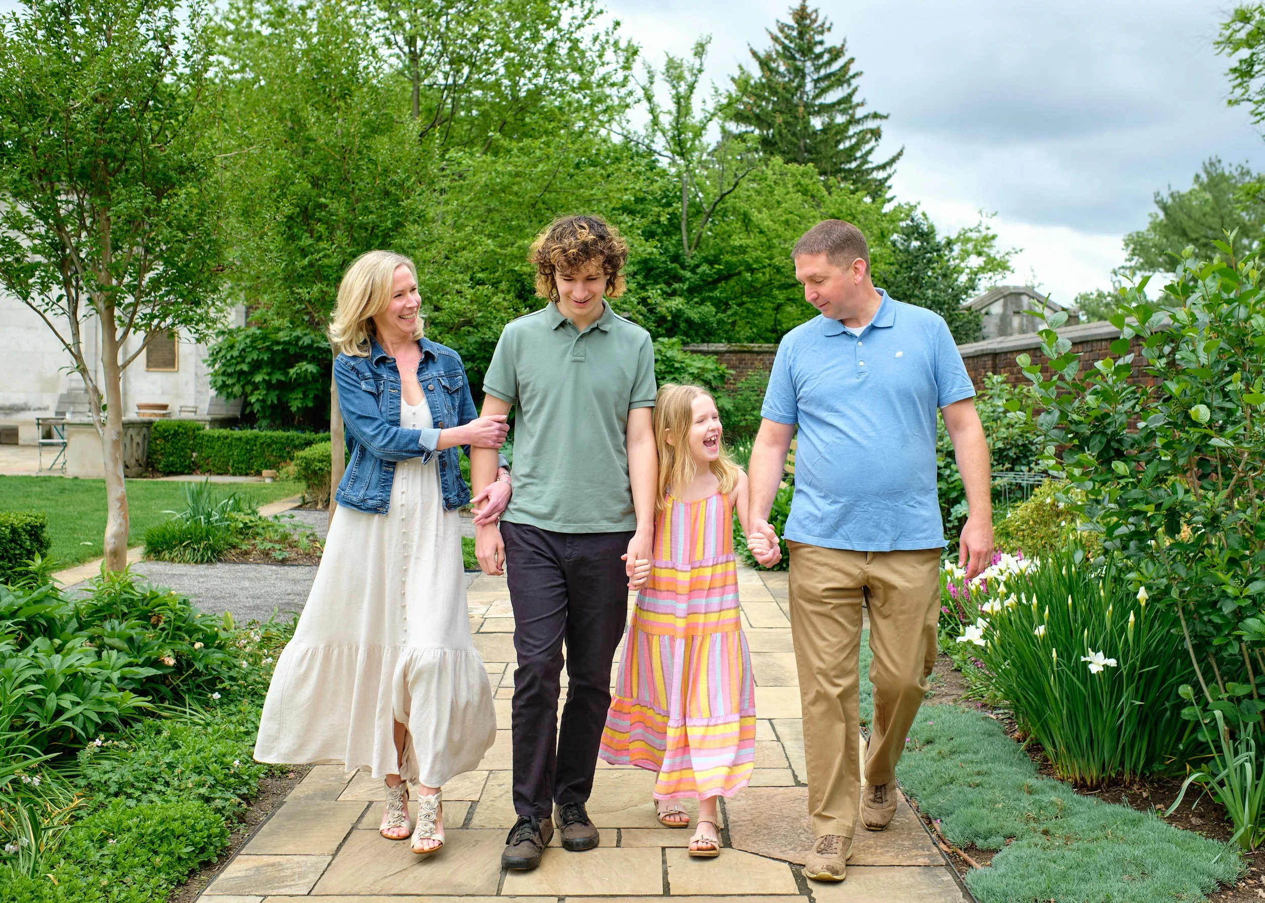 A family of five walking through a garden, holding hands, and smiling. The family includes two parents, a teenage boy, a young girl, and another young girl. The garden is lush with green trees, shrubs, and flowering plants, with a paved pathway.