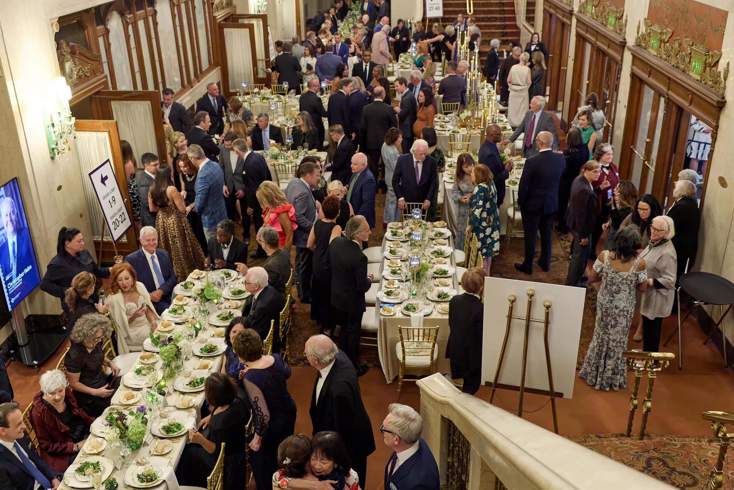 A large gathering of people at a formal event in a grand indoor venue with ornate decorations, long tables set with white tablecloths and floral centerpieces, and guests dressed in suits, dresses, and formal attire socializing and mingling.