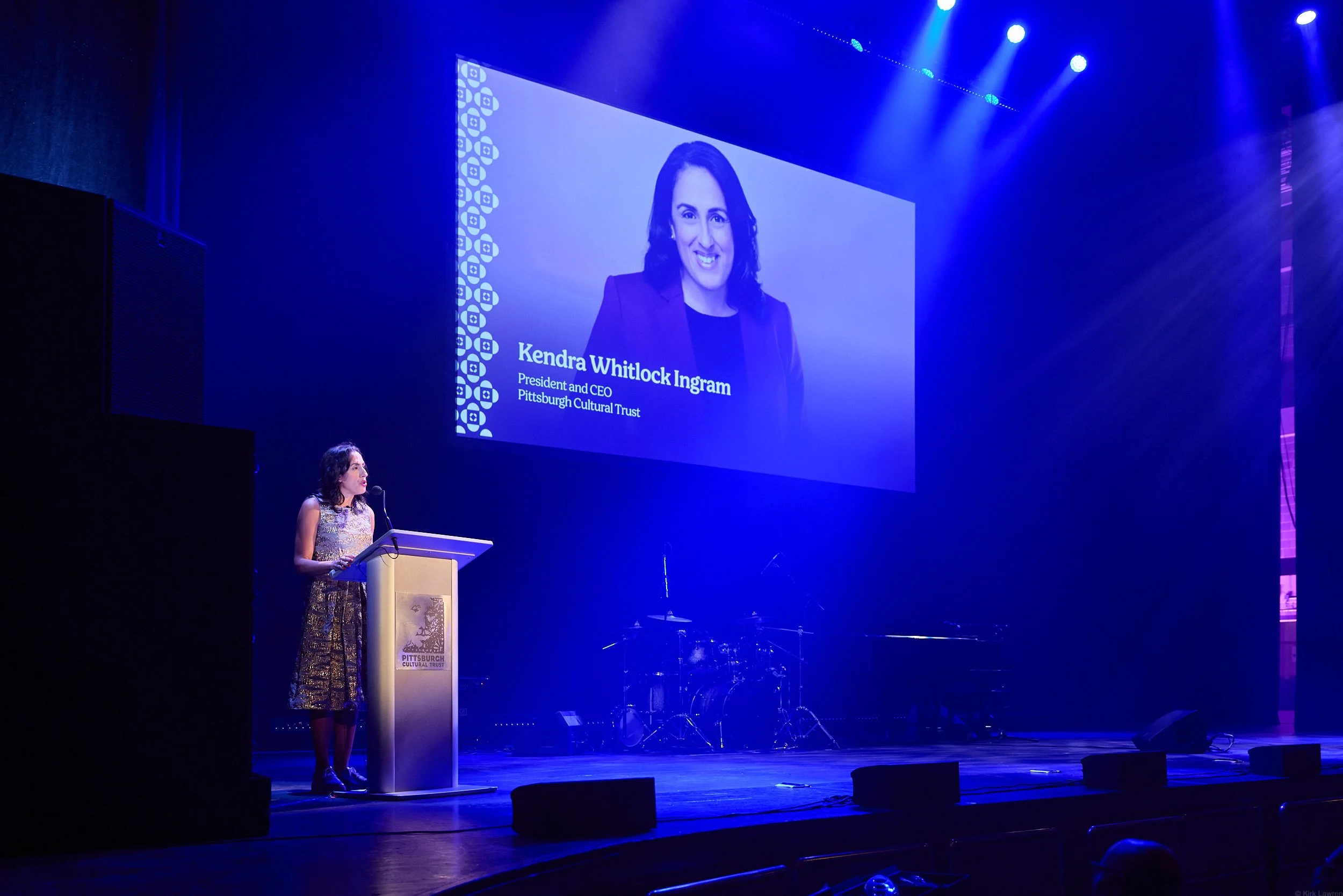 A woman stands at a podium on stage, speaking at an event. Behind her, a large screen displays a picture of a woman with dark hair, along with text that reads 'Kendra Whitlock Ingram, President and CEO, Pittsburgh Cultural Trust.' The stage is lit wi