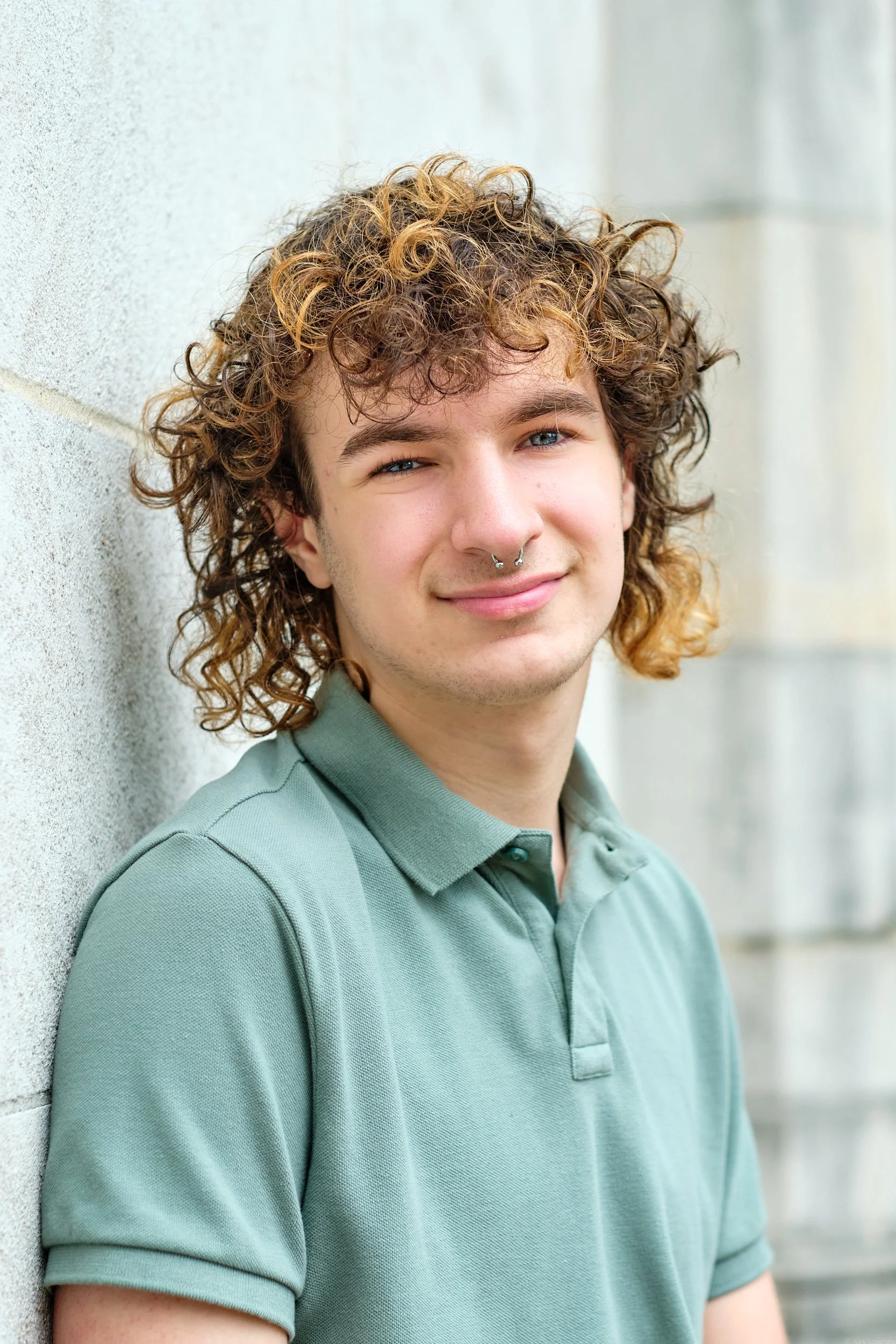 A young man with curly brown hair and blue eyes smiling, wearing a light green polo shirt, standing against a textured wall.