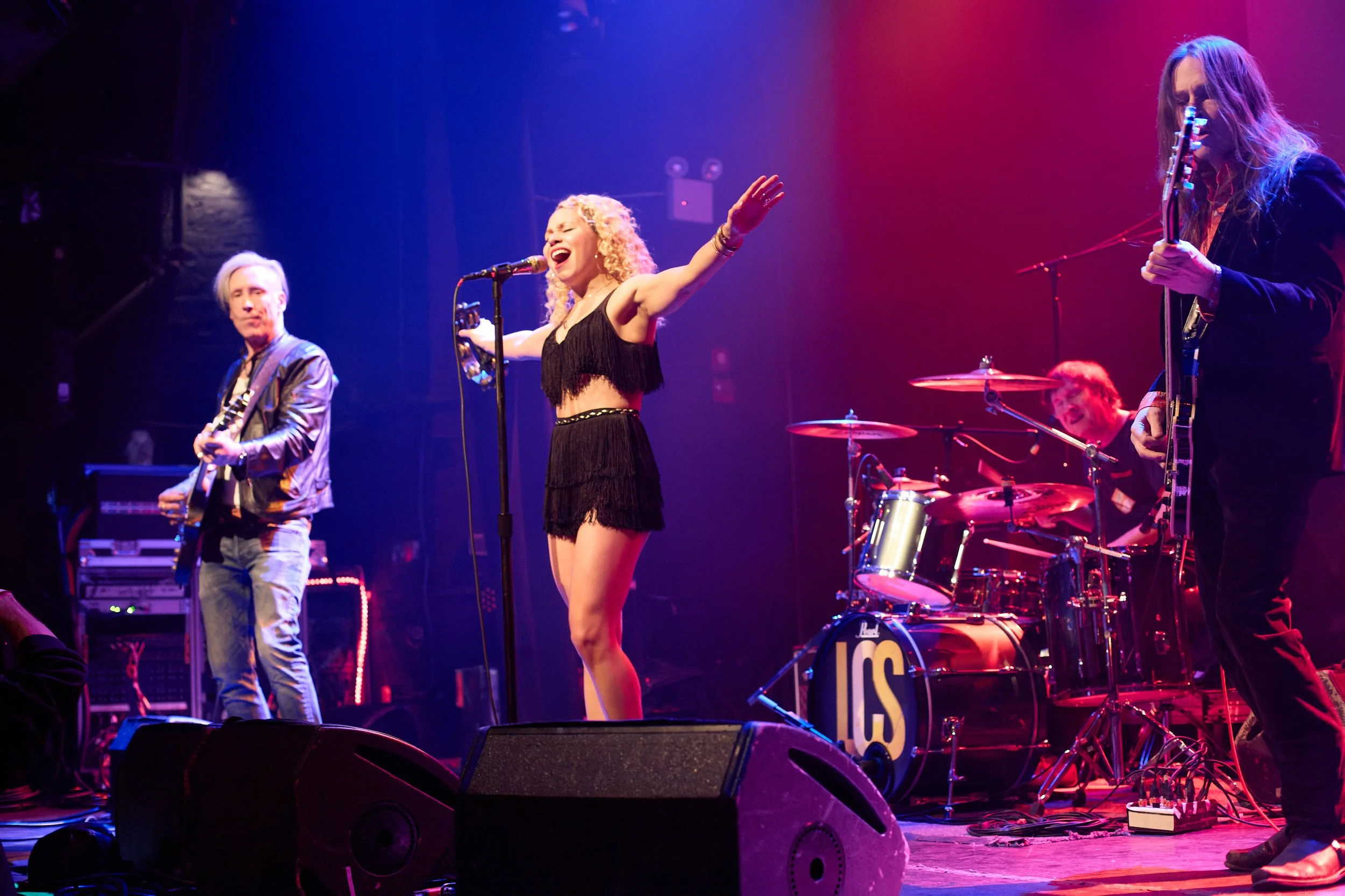 A band performing on stage with a female lead singer singing with her arms raised, guitarist on the right, bassist on the left, and drummer in the background under colorful stage lighting.