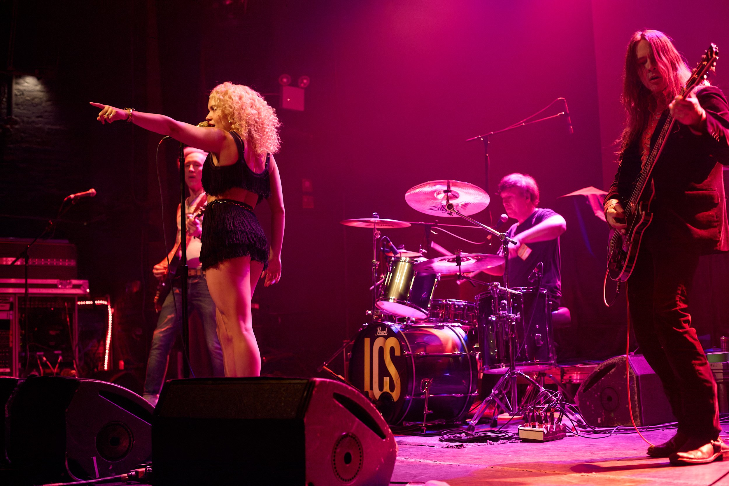 Band performing on stage with purple lighting, including a woman with curly blonde hair singing into a microphone, a woman playing guitar, and other musicians with drums and keyboard.