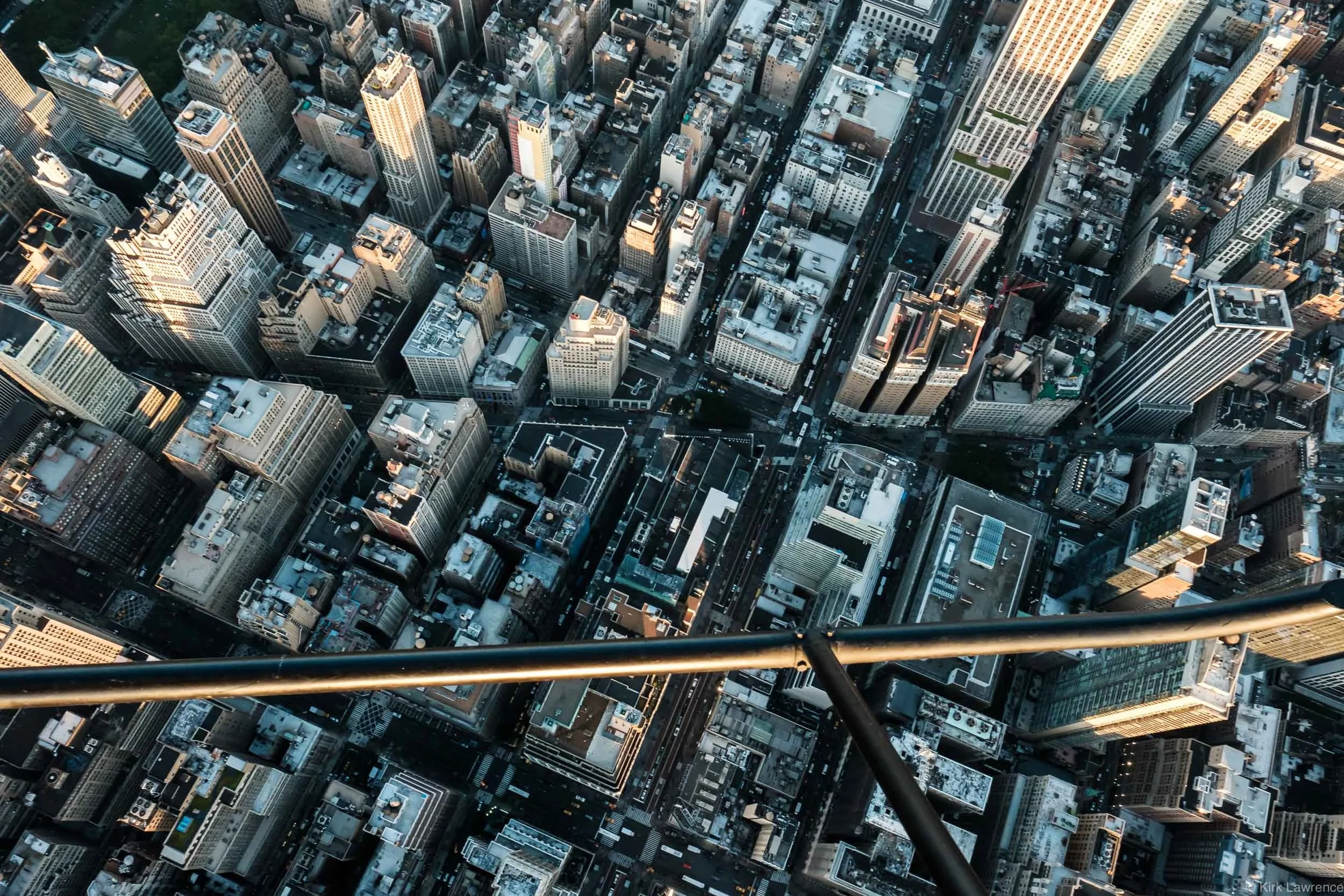 Aerial view of a dense cityscape with tall skyscrapers and streets below, taken from a helicopter with a landing skid visible in the foreground.