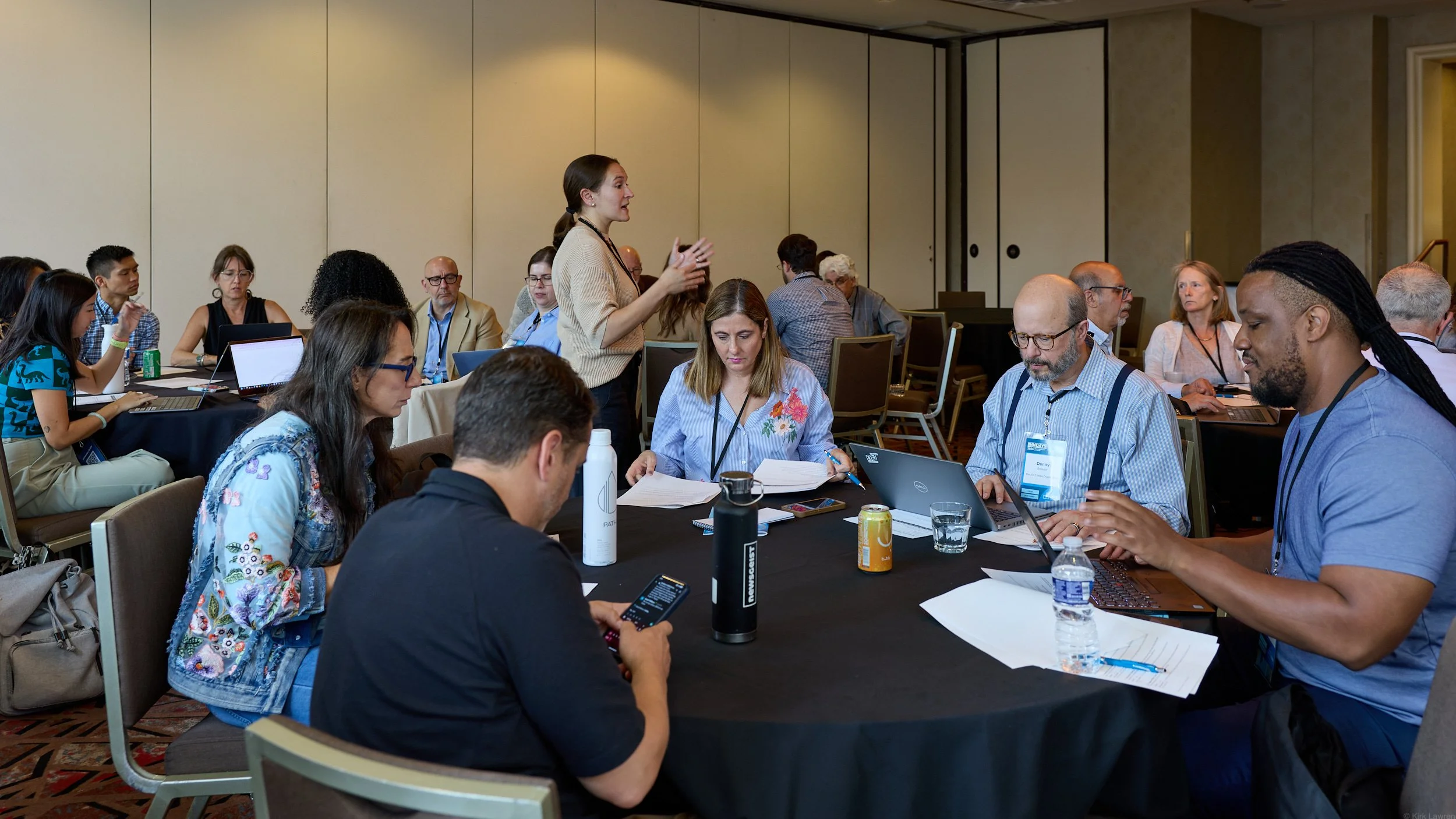 A diverse group of people sitting around a conference table in a meeting room, with a woman standing and speaking to the group.