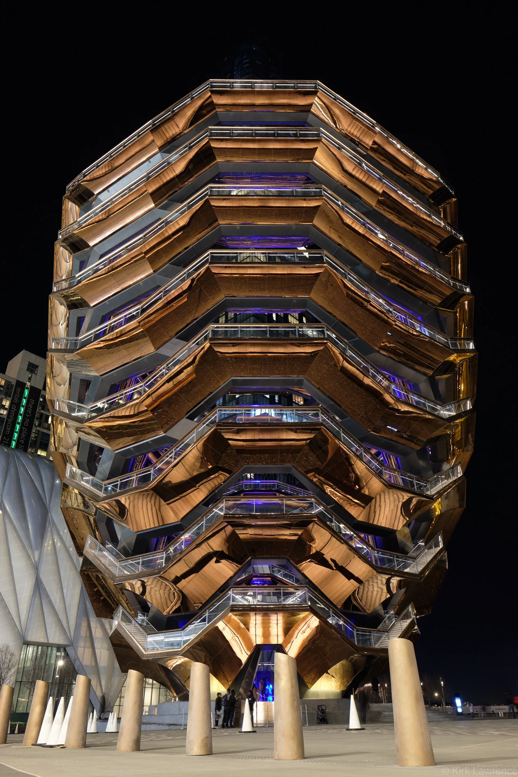 Night view of the Vessel, a copper-colored, honeycomb-like architectural structure in New York City with illuminated led lights and glass railings, surrounded by concrete pillars and a few people at its base.