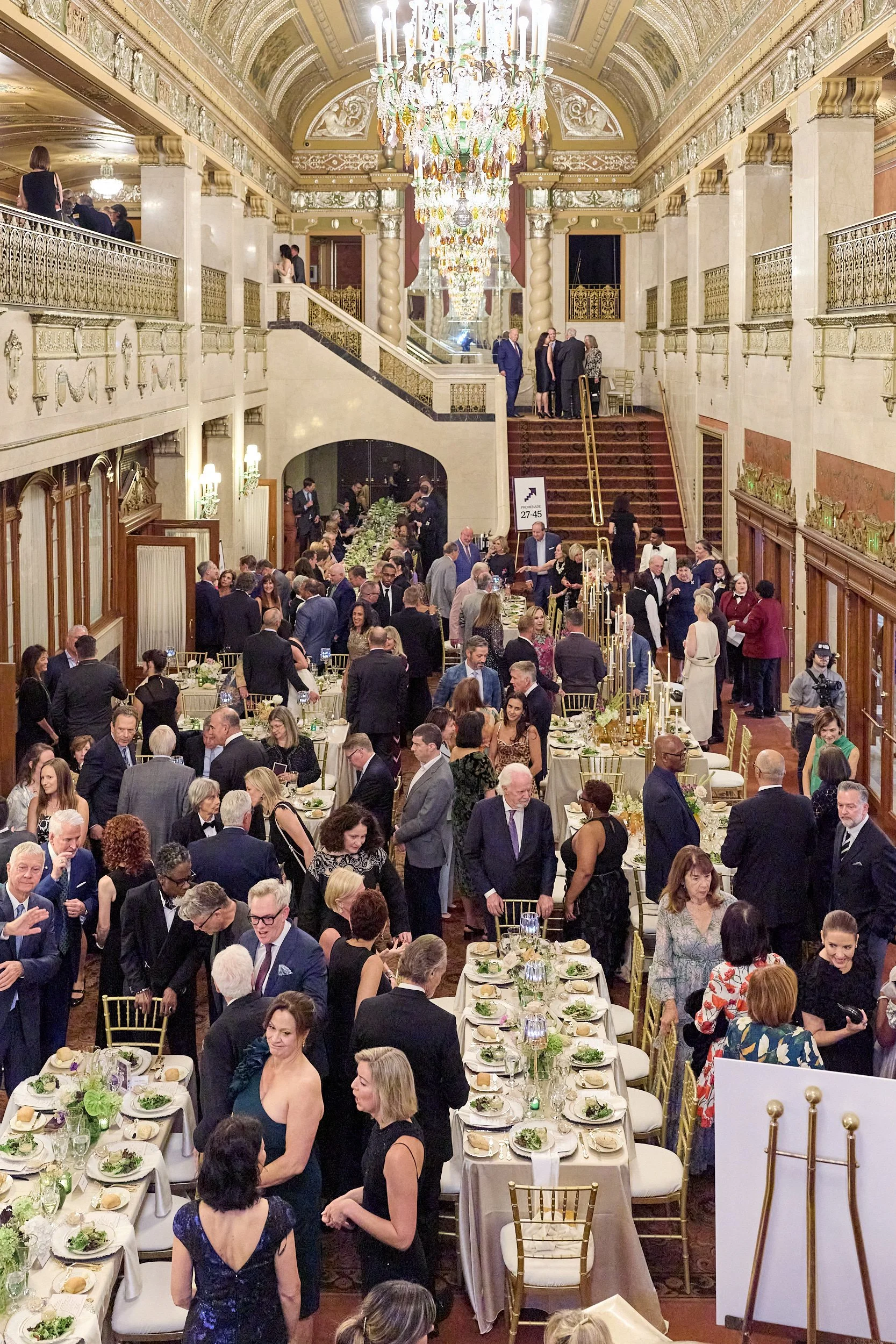 Elegant indoor banquet with guests socializing, sitting at decorated tables, and standing on a staircase in a grand hall with chandeliers and ornate decor.