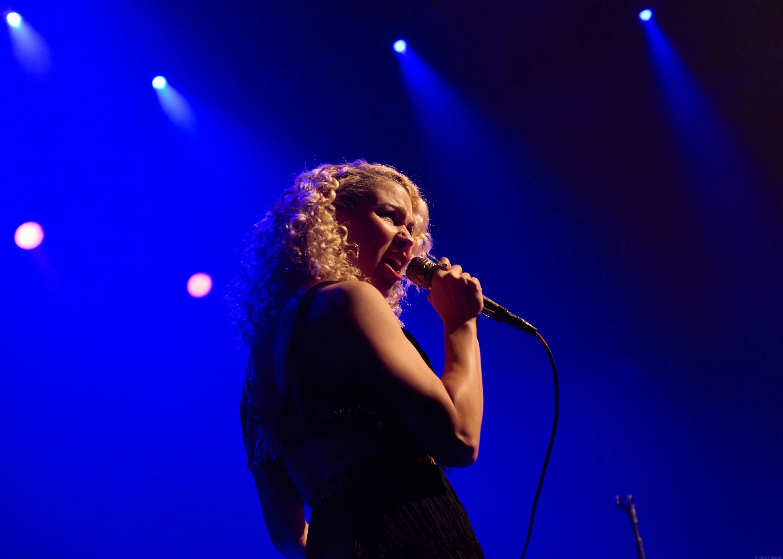 A woman with curly blonde hair singing into a microphone on stage, illuminated by blue stage lights.
