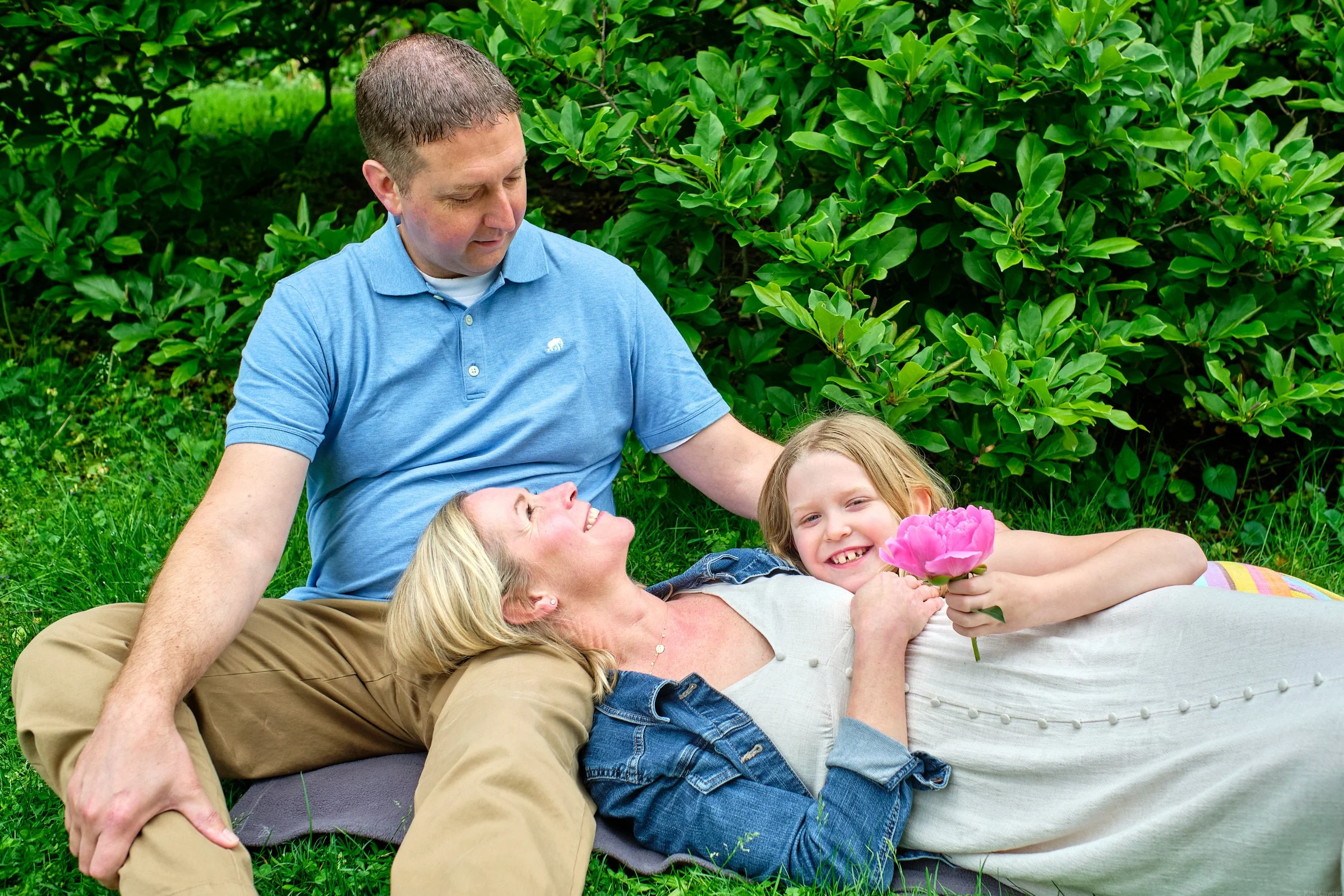 A family of four sitting outdoors on grass, with green bushes in the background. The mother is lying down with her head on the father's lap, smiling and holding a pink flower. The father is sitting upright, looking at his wife. The two children are l