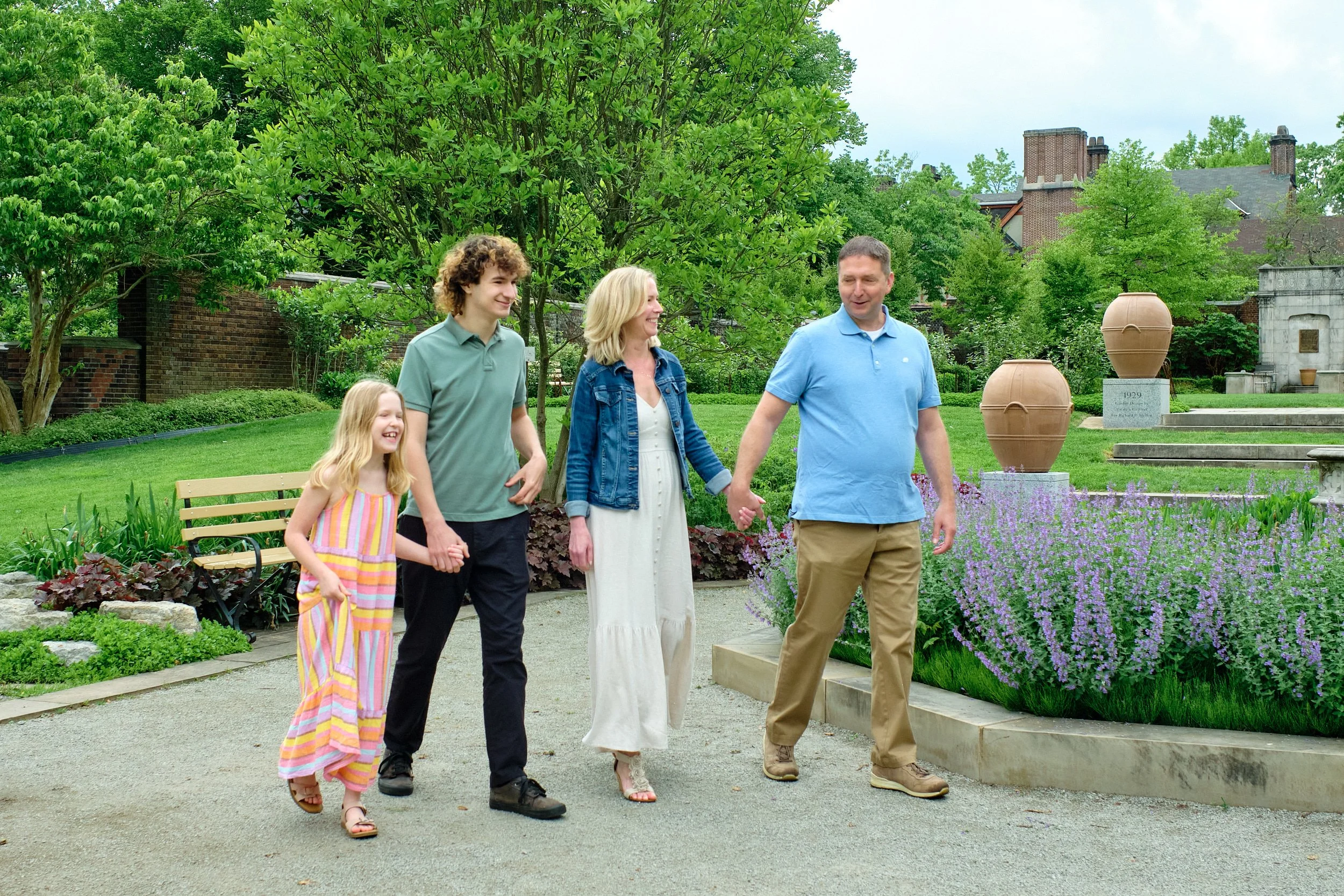 A family of five walking in a lush, green garden with colorful flowers and large decorative pots. The family appears happy and relaxed, enjoying a pleasant day outdoors.