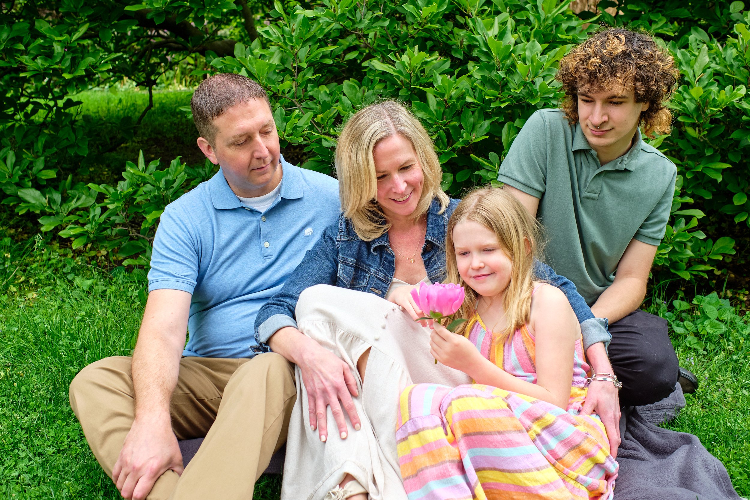 A family of five sitting on grass and surrounded by green bushes. The mother and father sit with two daughters, while a teenage boy sits next to them. The daughters are holding a pink flower, and everyone is smiling.