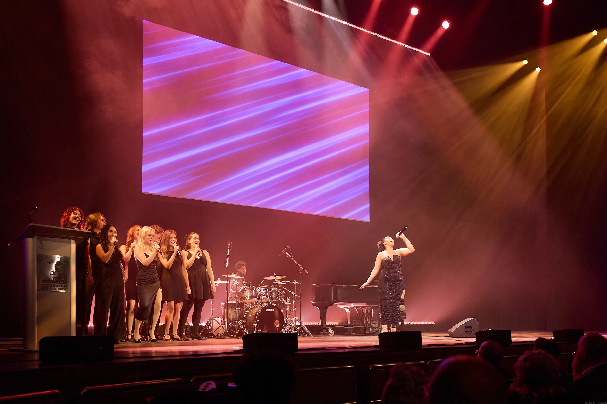A group of women standing on a stage, singing into microphones, with a drummer and drinks in the background. A woman in a black dress stands apart, singing into a microphone. There is a large screen with purple and pink lights in the background, and 
