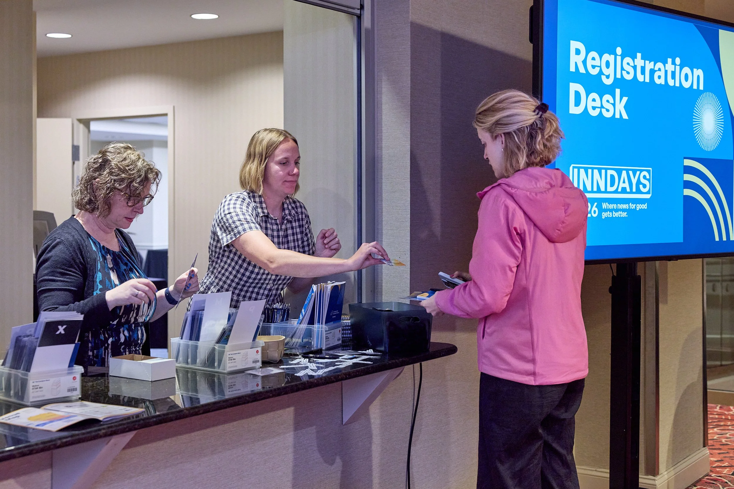 A woman in a pink jacket is standing at a registration desk, handing over a card or paper to a staff member. The staff member is seated behind the counter, assisting her, and another staff member is nearby, looking at papers. Behind the desk, a large