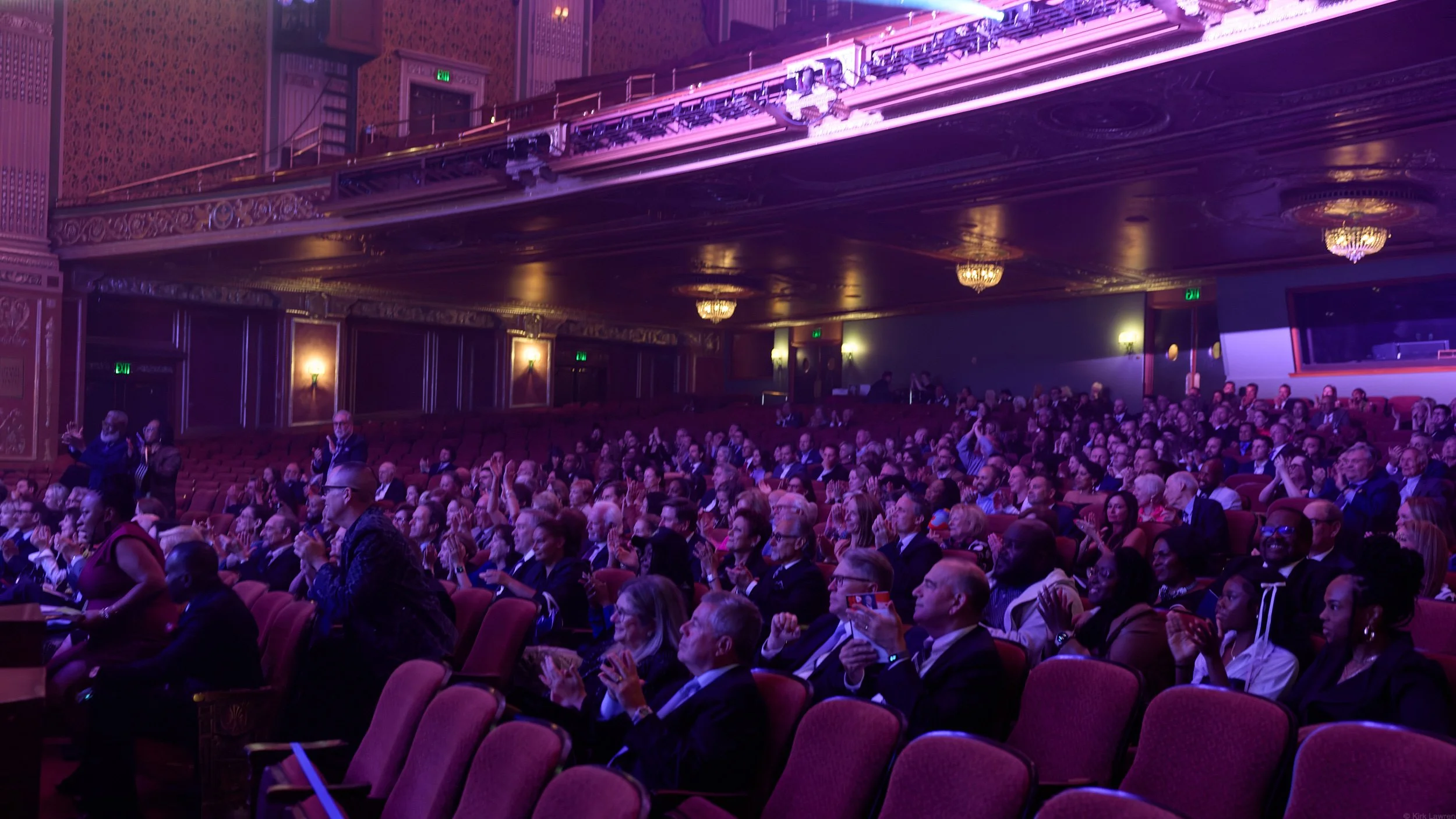 Audience gathered in a theater with ornate ceiling and purple lighting, clapping for a performance.