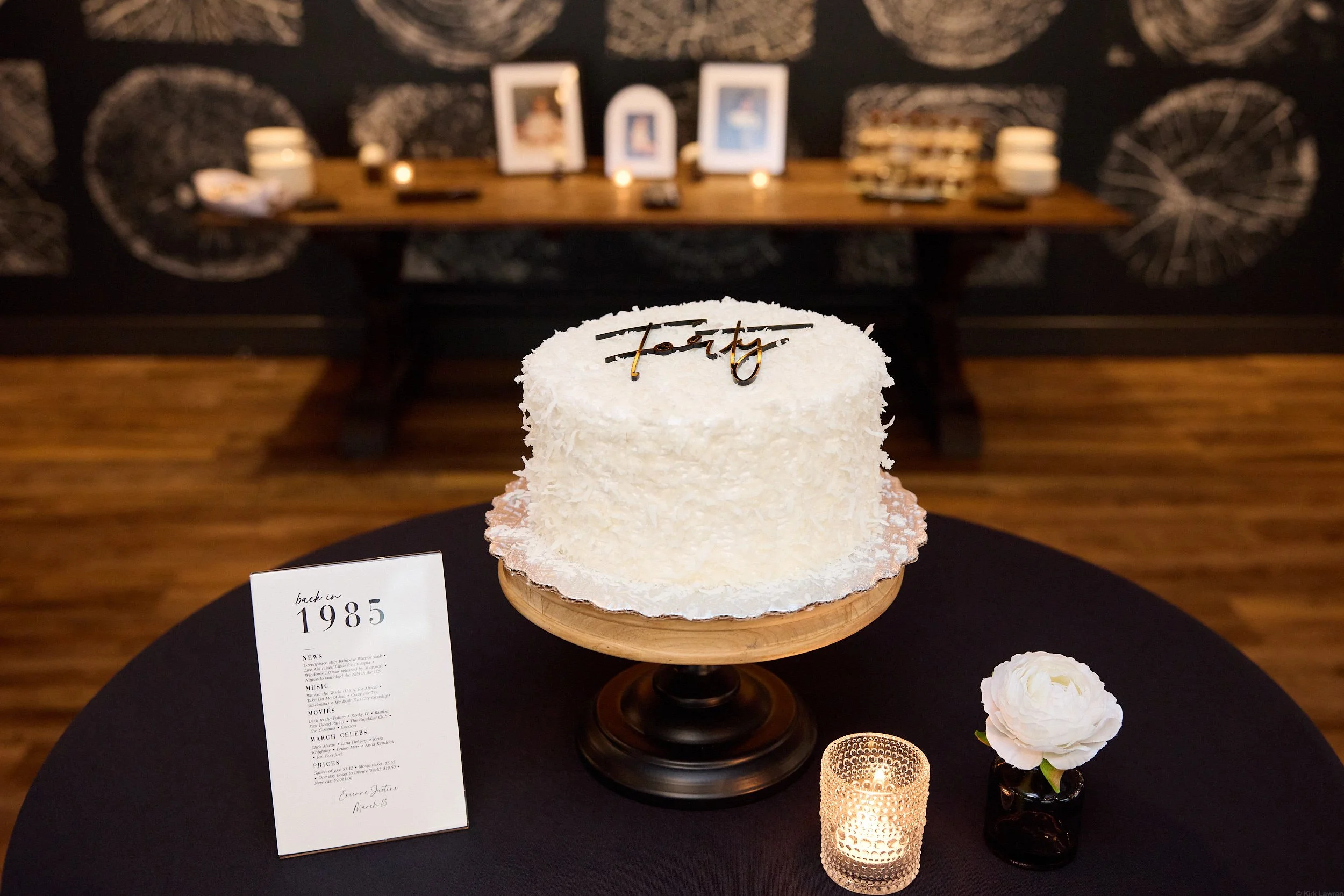 A white coconut cake with a gold 'Party' topper, sitting on a black table surrounded by a white flower in a black vase, a candle holder, and an informational card, with a dark wall featuring artistic circular patterns and framed pictures in the backg