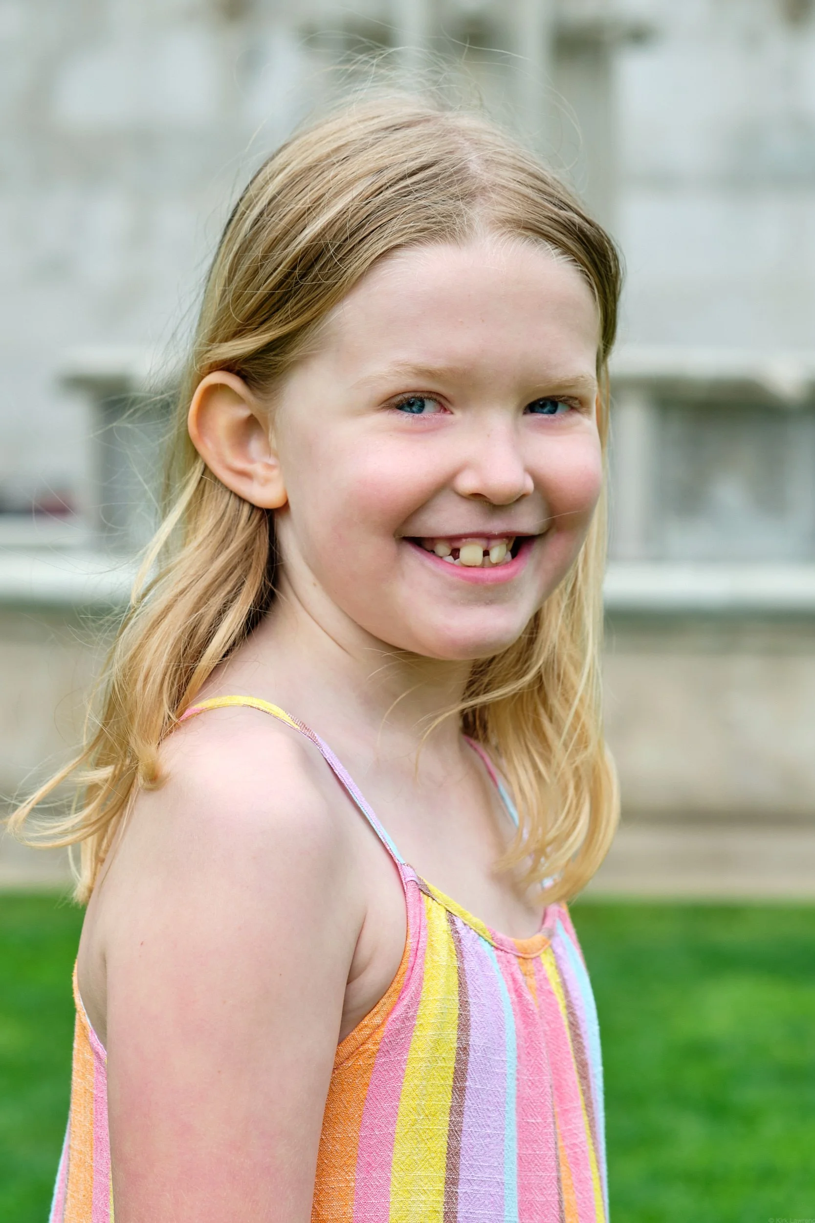 A young girl with long blonde hair and blue eyes smiling outdoors with blurred greenery and a white structure in the background, wearing a colorful striped sundress.