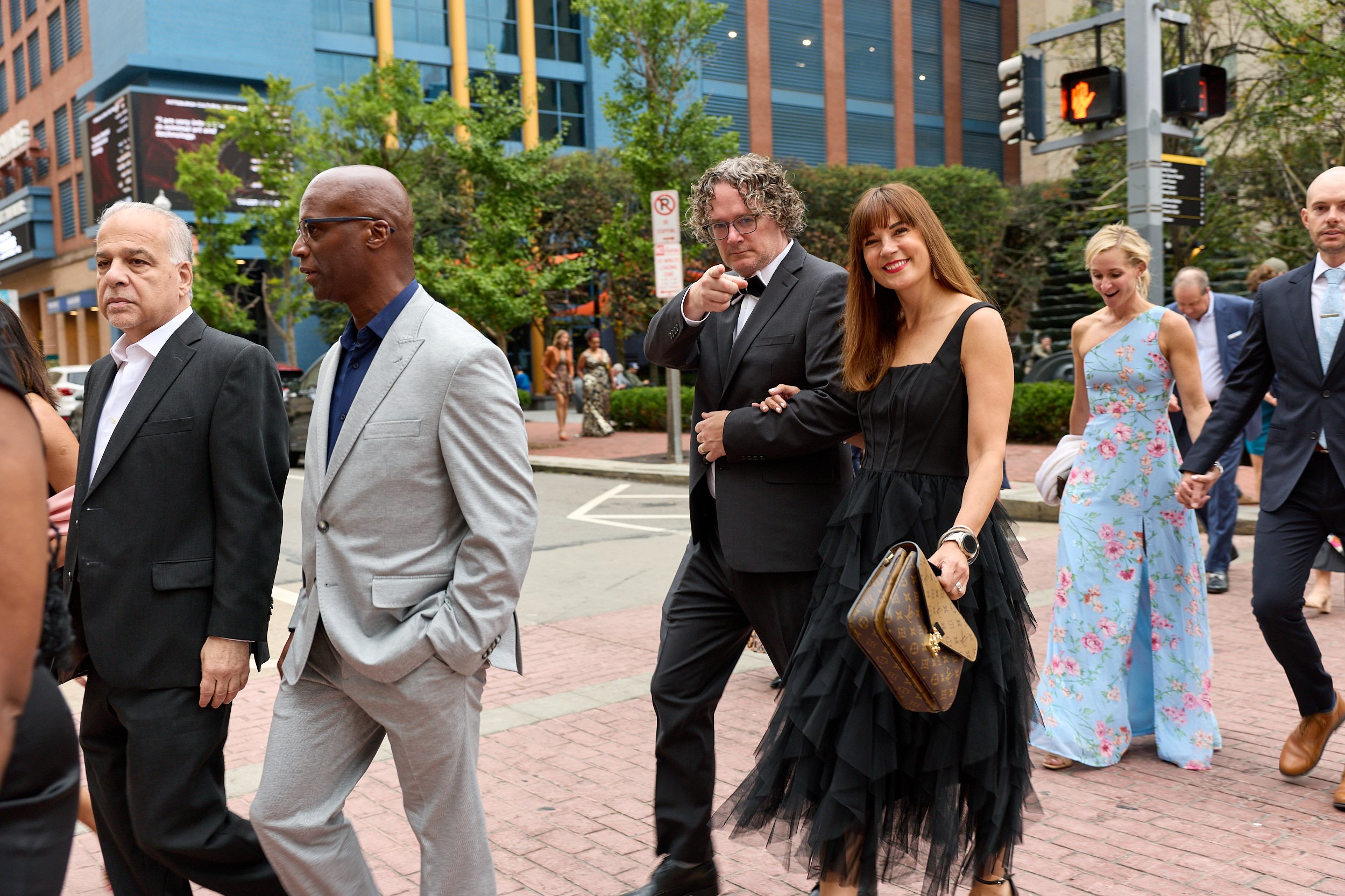Group of people dressed in formal and semi-formal attire walking across city street with modern buildings, traffic lights, and trees in the background.