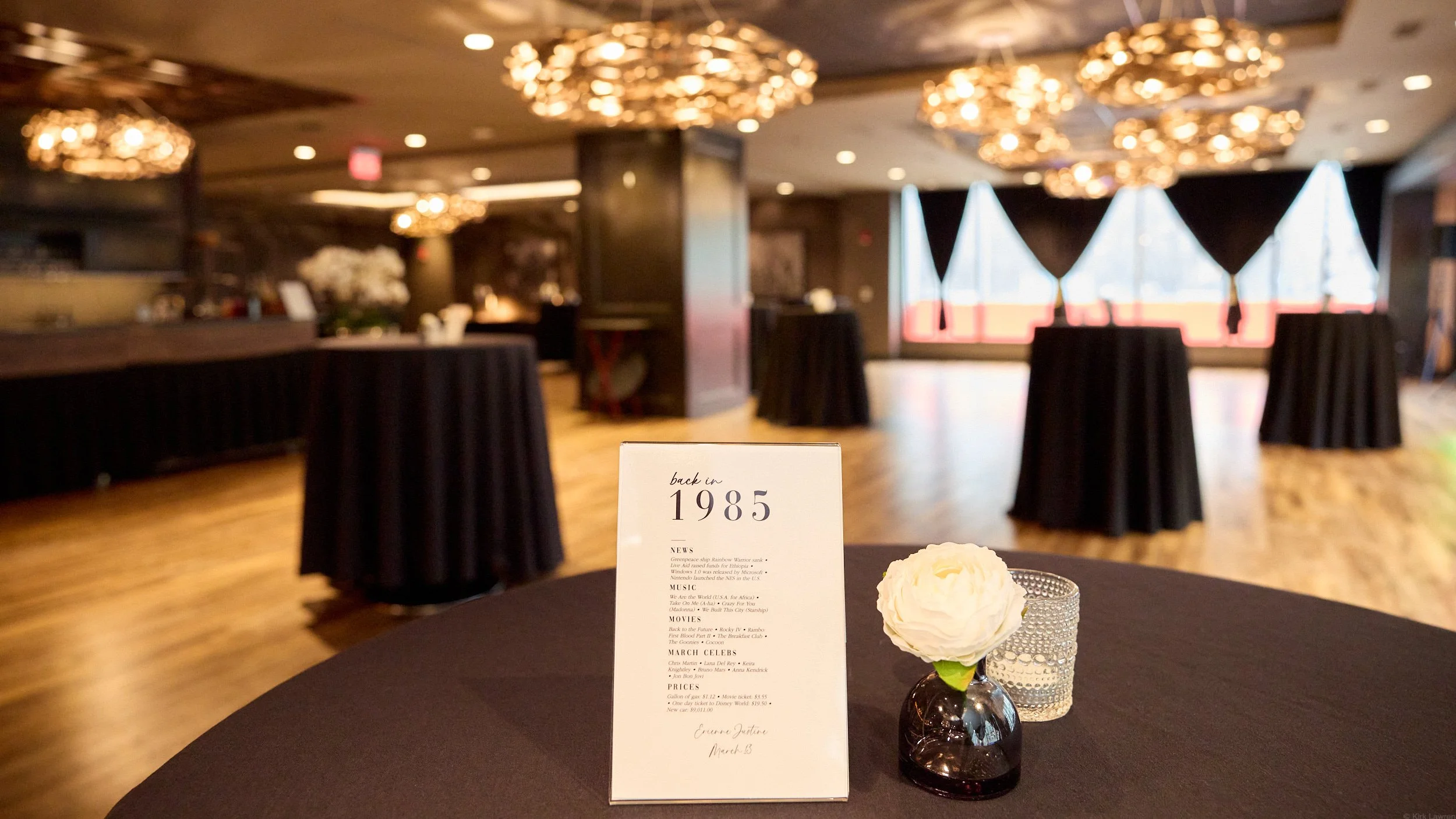 Interior of a banquet or event space with round tables covered in black tablecloths and floral centerpieces. The room features modern chandeliers and large windows with dark curtains, and a small sign on one table indicates the theme is "back in 1985