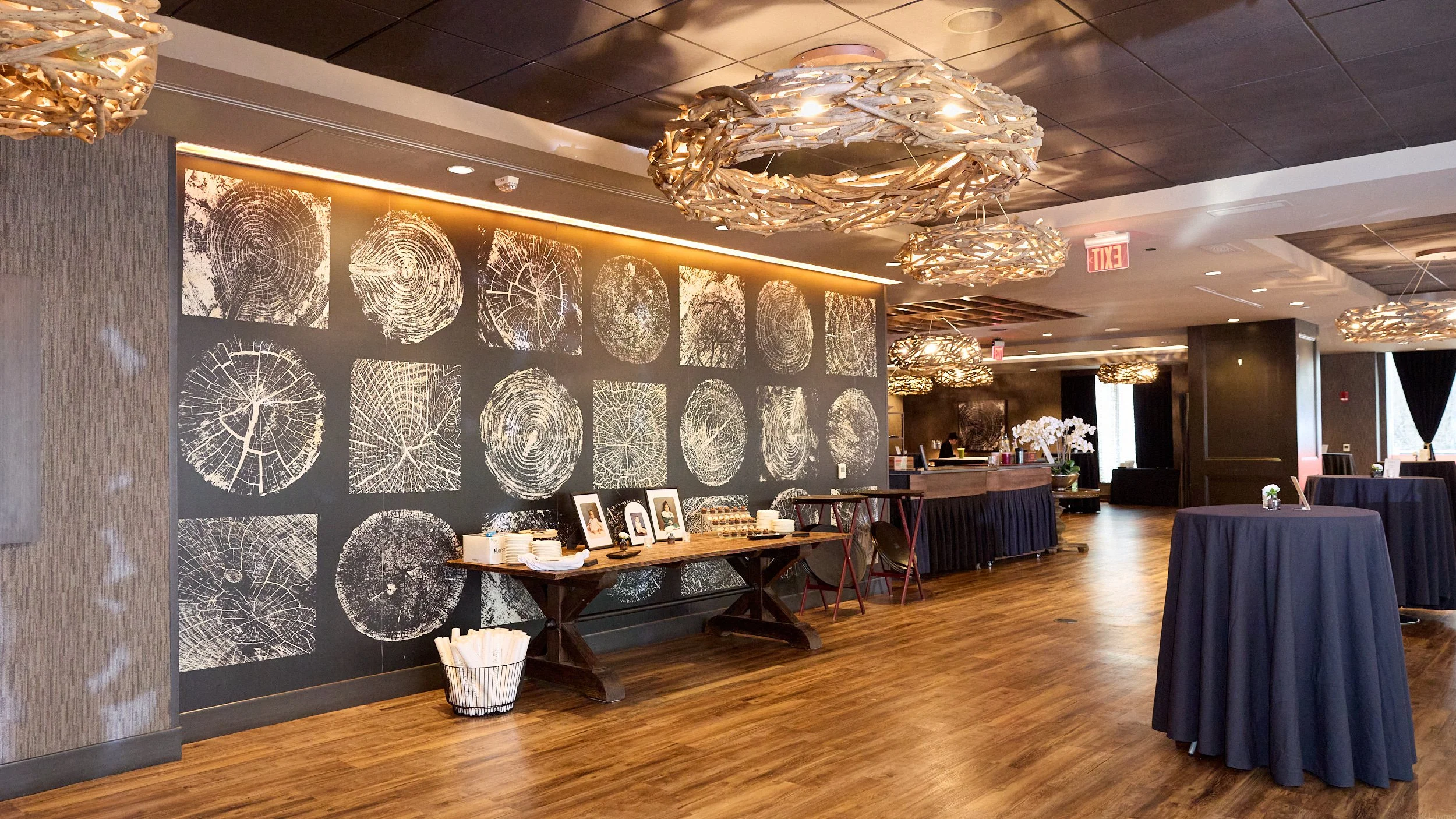 Interior of a modern event space with artistic wood grain wall decor, hanging light fixtures made of intertwined wood pieces, round tables with dark tablecloths, and a buffet area with white plates and framed pictures.