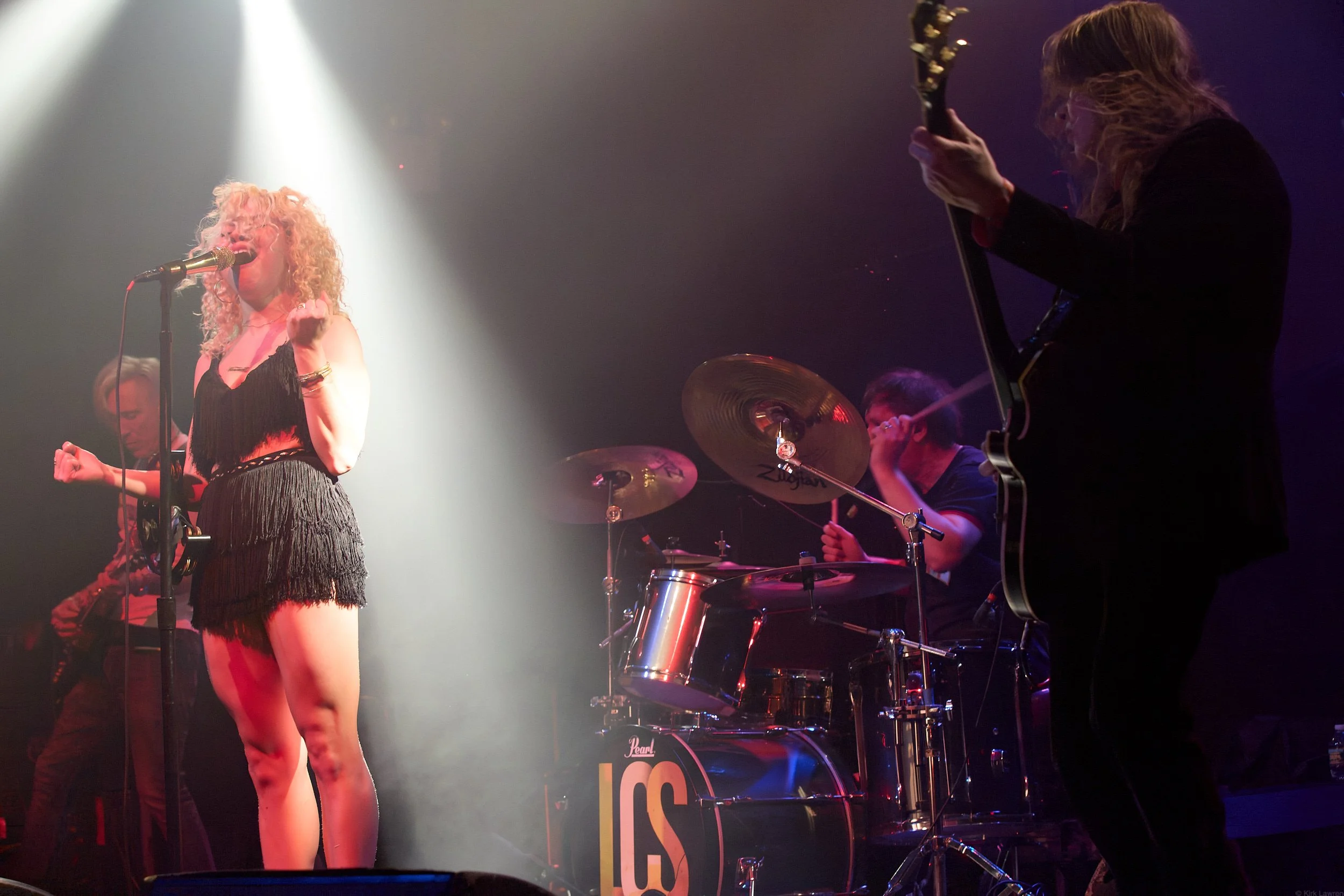 A woman with curly blonde hair singing into a microphone on stage, accompanied by a drummer and guitarist, with stage lights focused on her.
