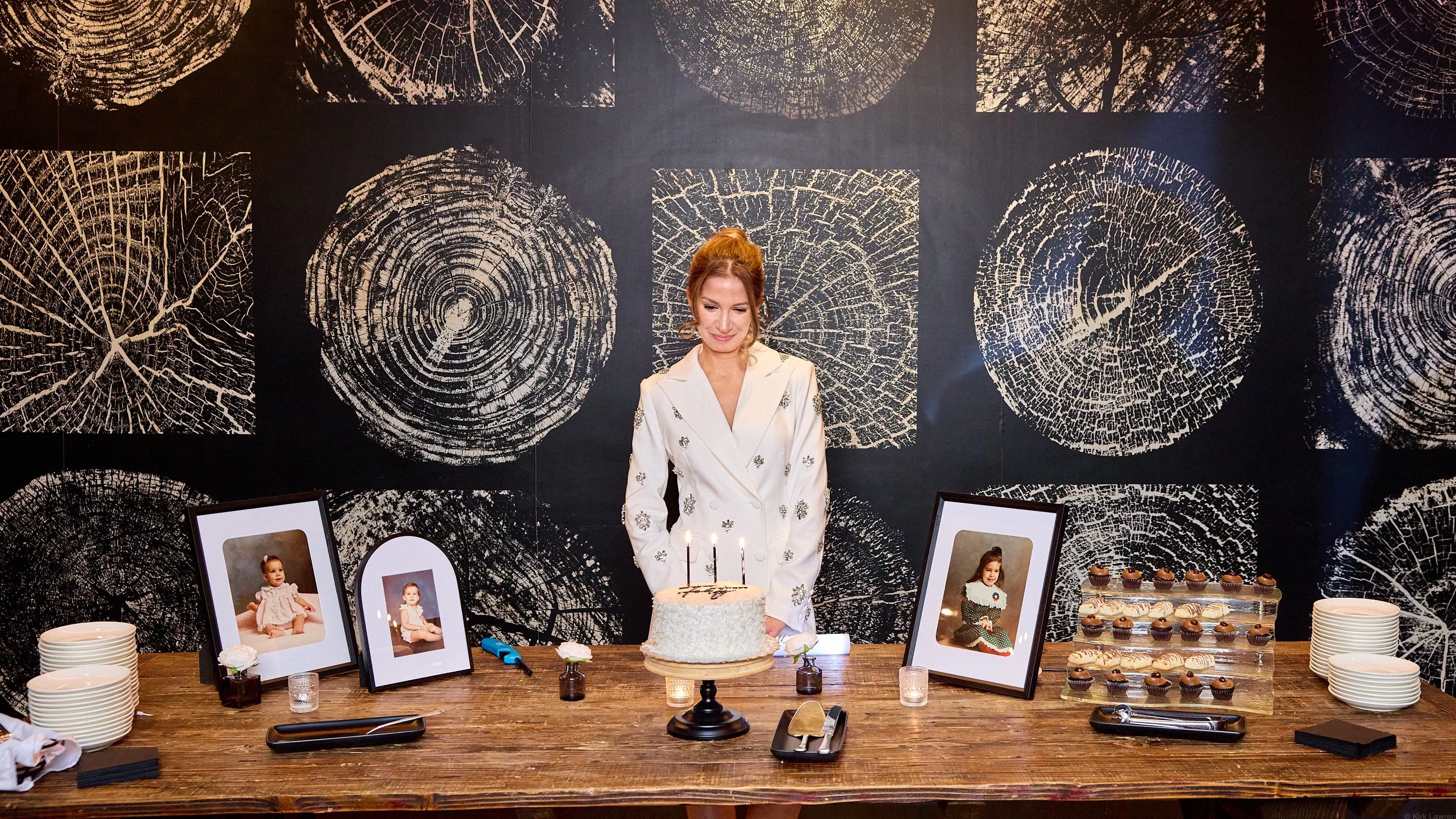 A woman in a white dress standing behind a birthday cake with lit candles, surrounded by framed photographs of a young girl, cupcakes, and plates on a wooden table, with black and white tree ring wall art in the background.
