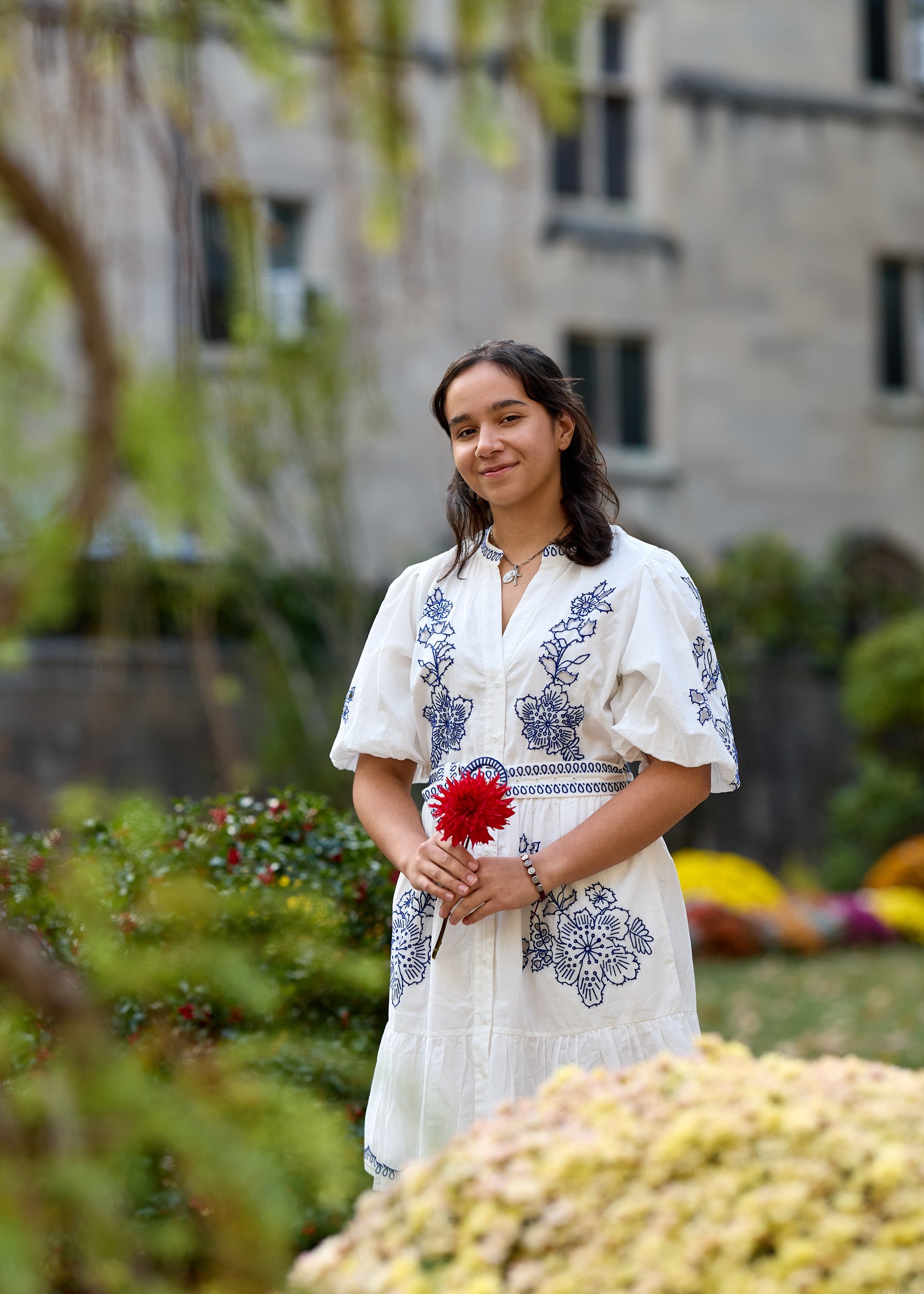 A young woman with shoulder-length dark hair, wearing a white dress with blue embroidery, stands outdoors holding a red flower. She is smiling slightly and appears to be in a garden with colorful flowers and a blurred building in the background.