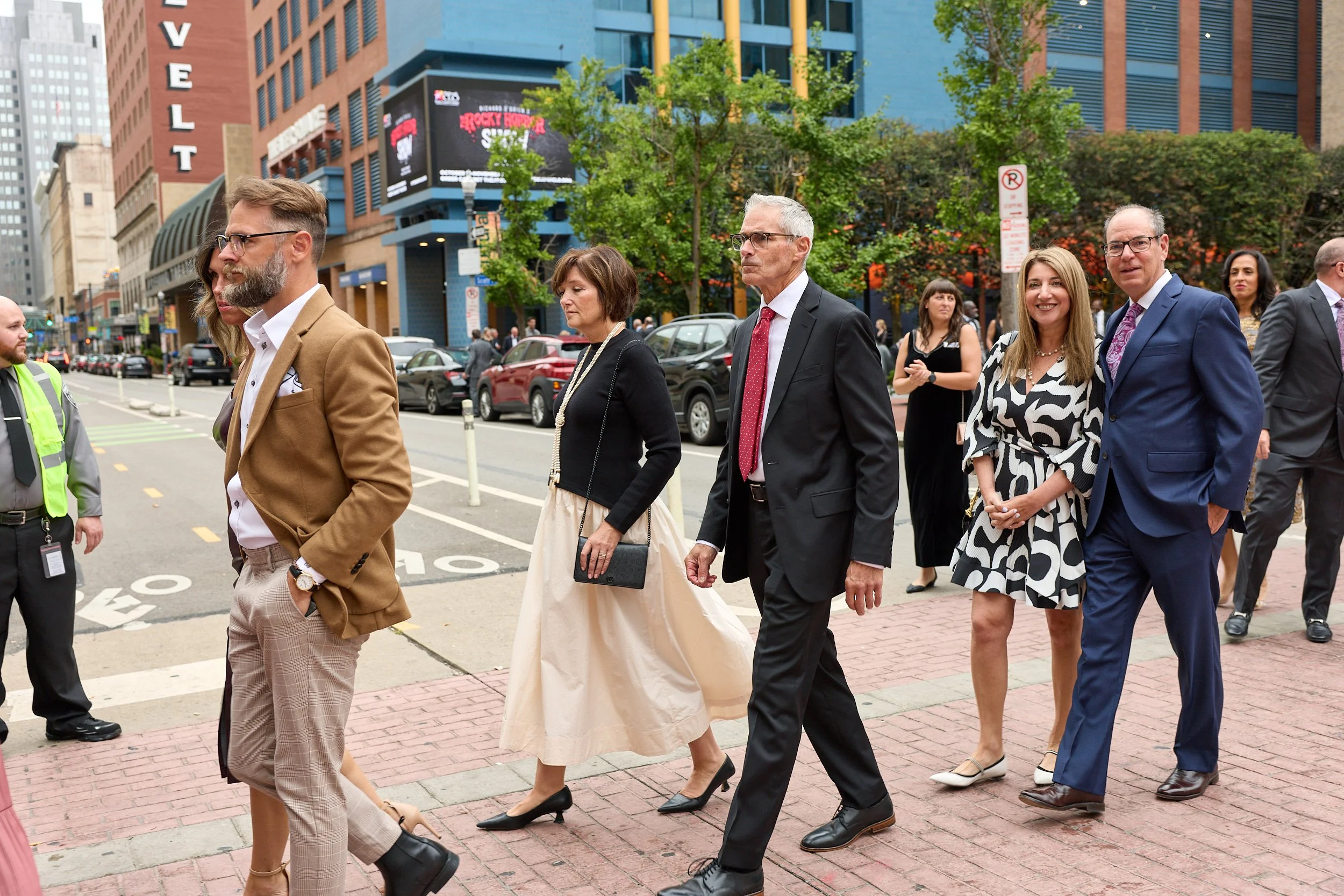 A group of men and women dressed in business and fashionable attire walk across a city street in front of tall buildings and trees during daytime.