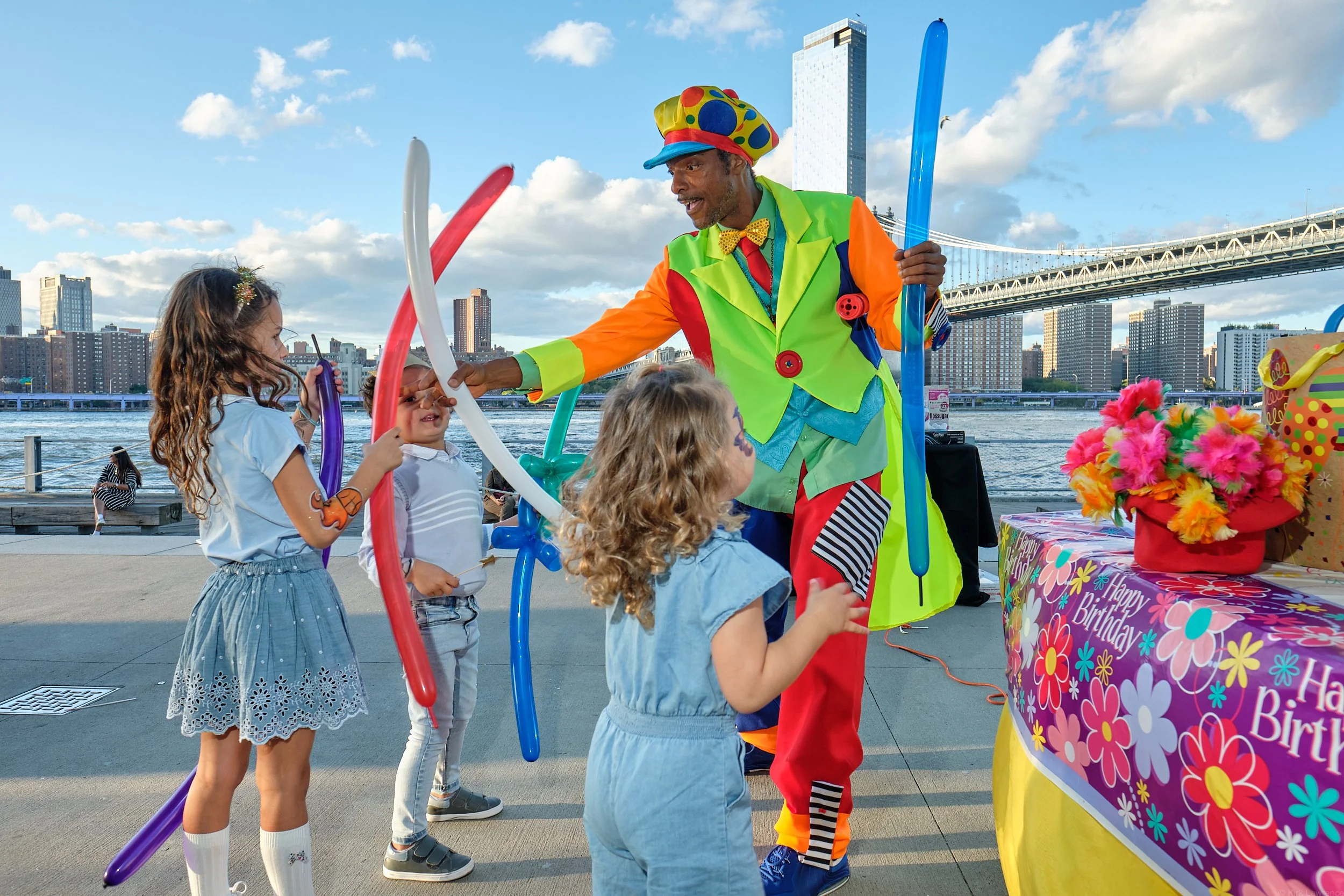 A clown dressed in bright colorful costume performing balloon art for children near a waterfront with a New York City skyline and the Manhattan Bridge in the background.
