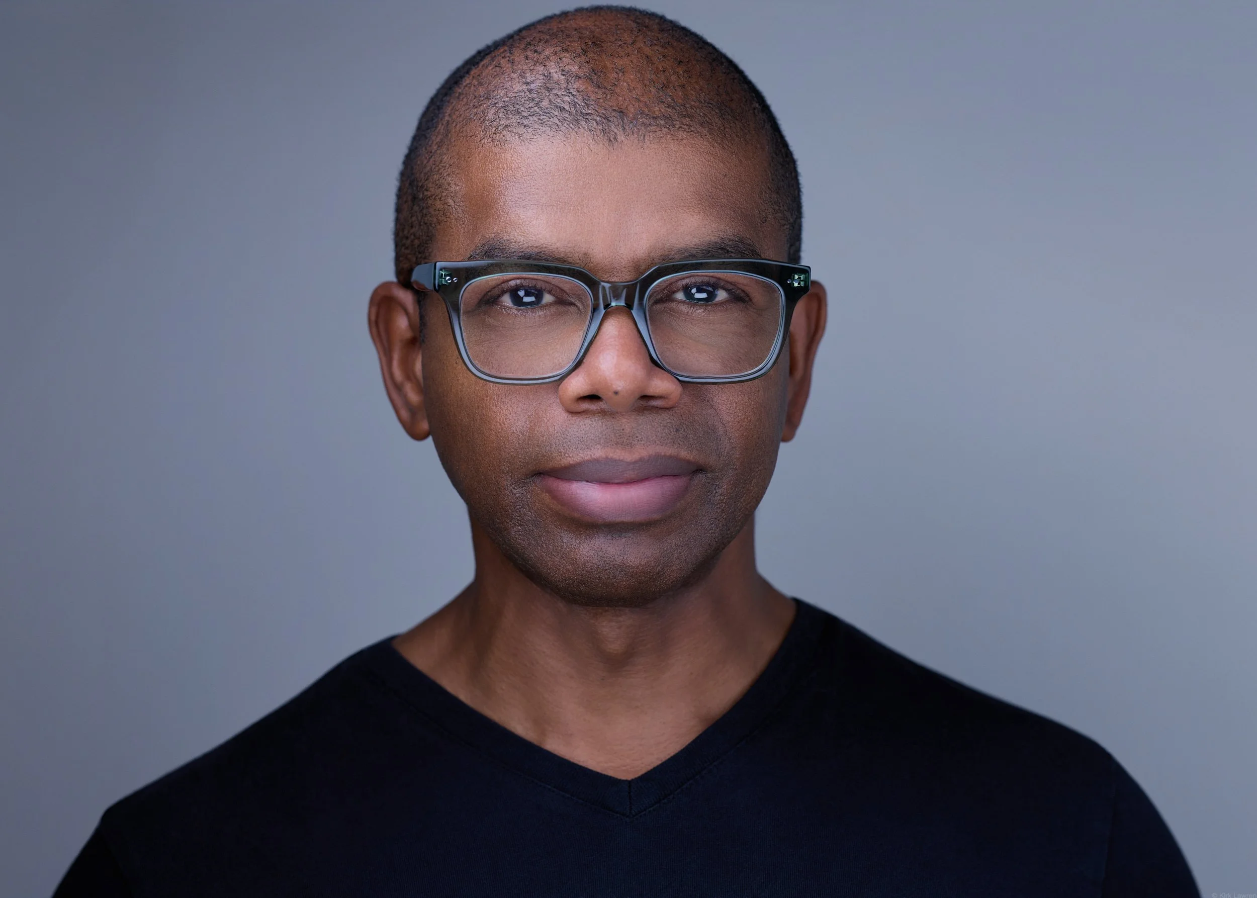 Close-up portrait of a man with short hair, wearing black-rimmed glasses and a black shirt, against a plain gray background.