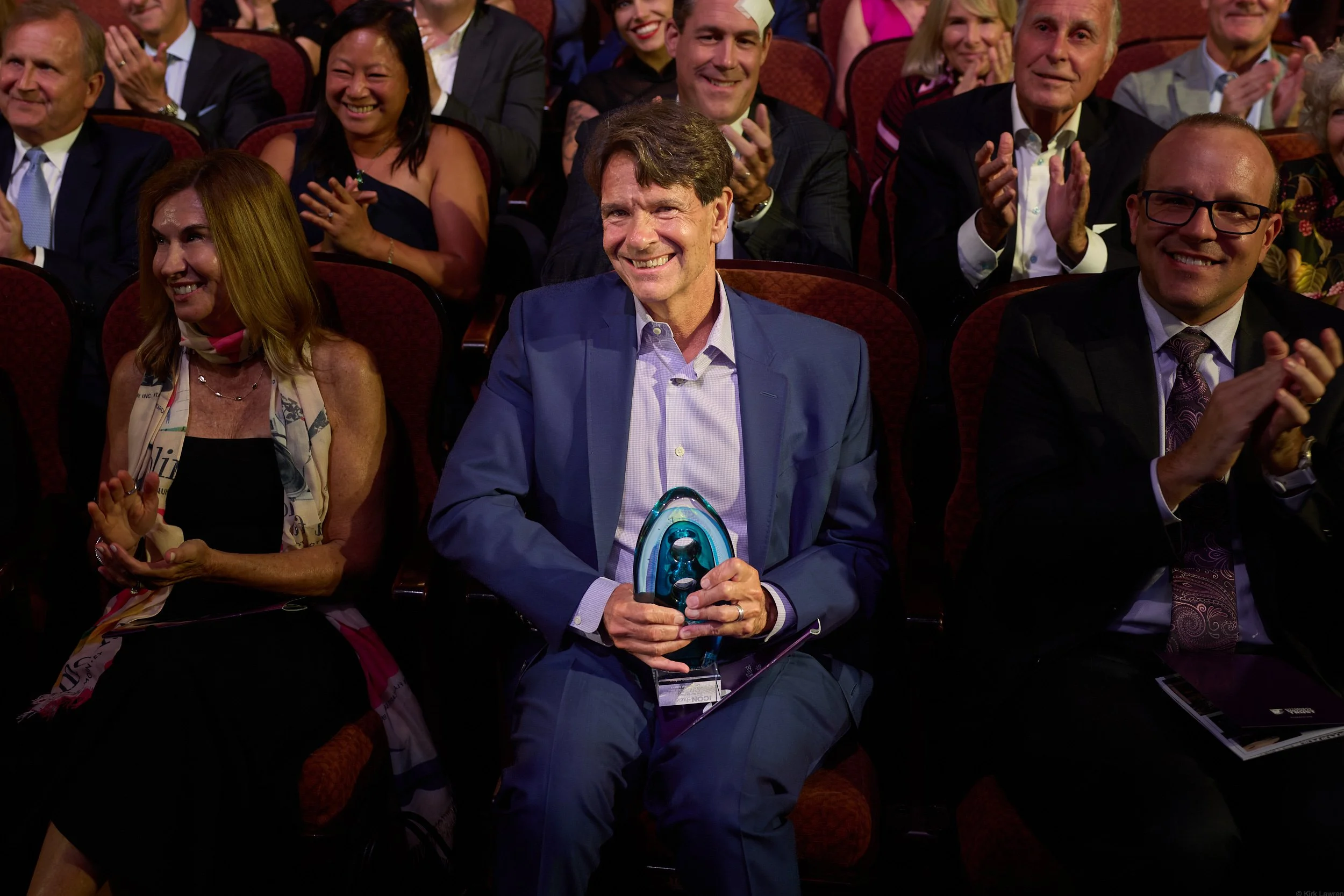 A group of people dressed in formal attire, seated in an auditorium, applauding a speaker or performer. The man in the center, wearing a blue suit, is smiling and holding an award or trophy.