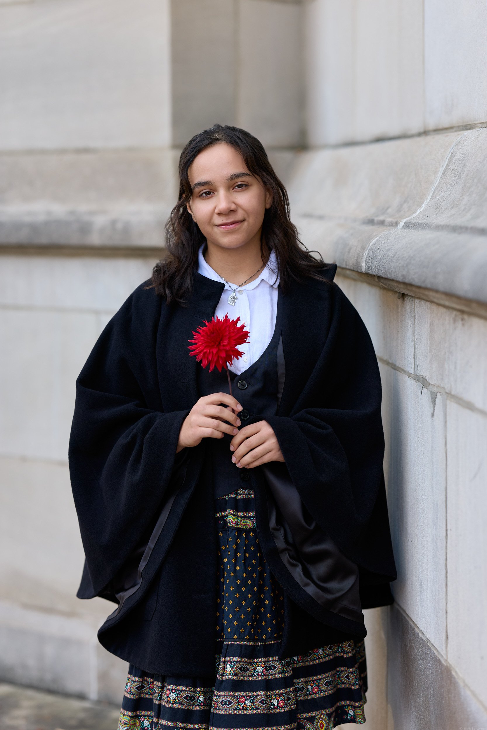 A young woman with dark wavy hair holding a red flower, standing against a beige stone wall, wearing a white shirt, black jacket, and a dress with colorful patterns.