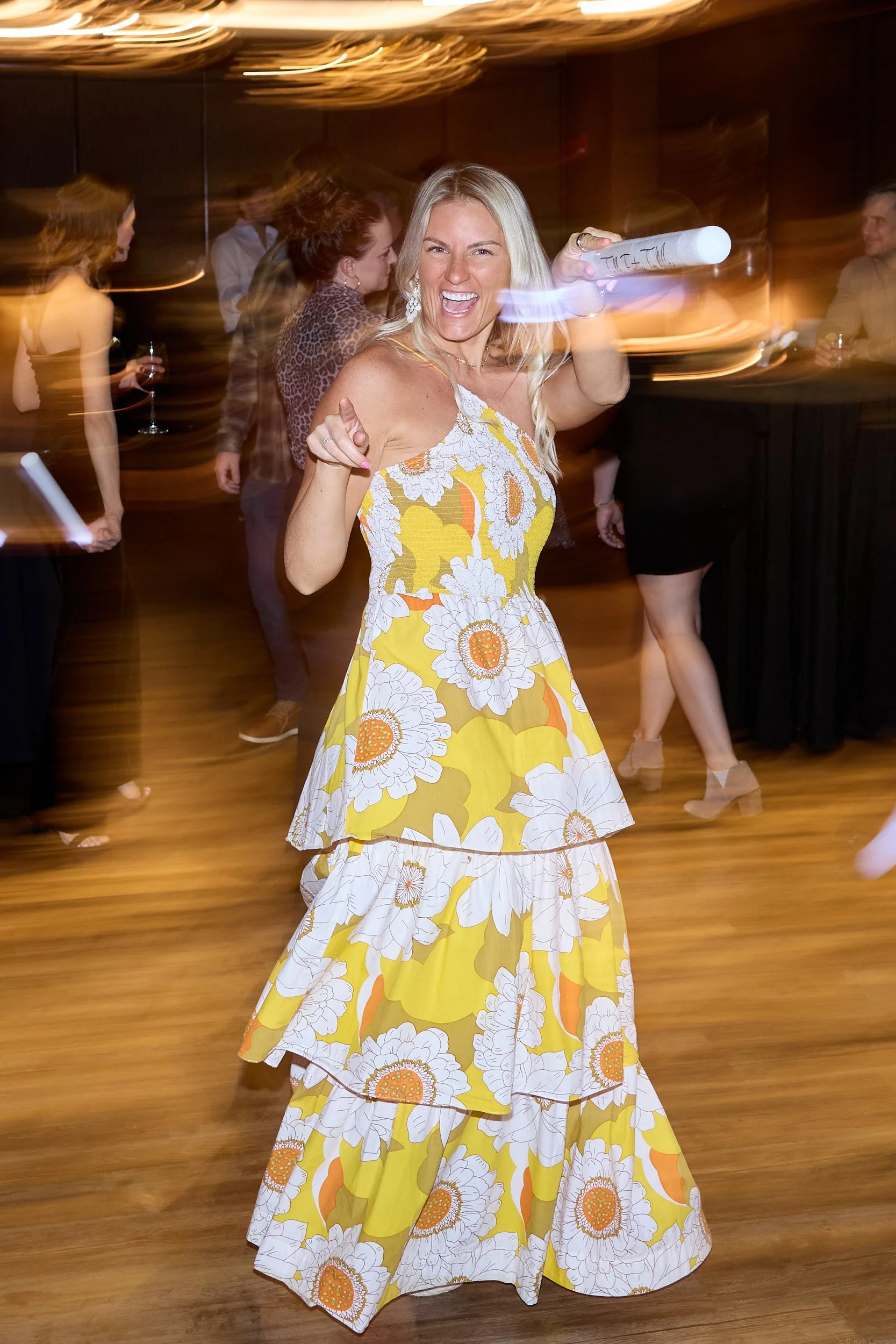 A woman in a long yellow and white floral dress holding a rolled up piece of paper, smiling and pointing towards the camera at an indoor event with blurred background of other people.
