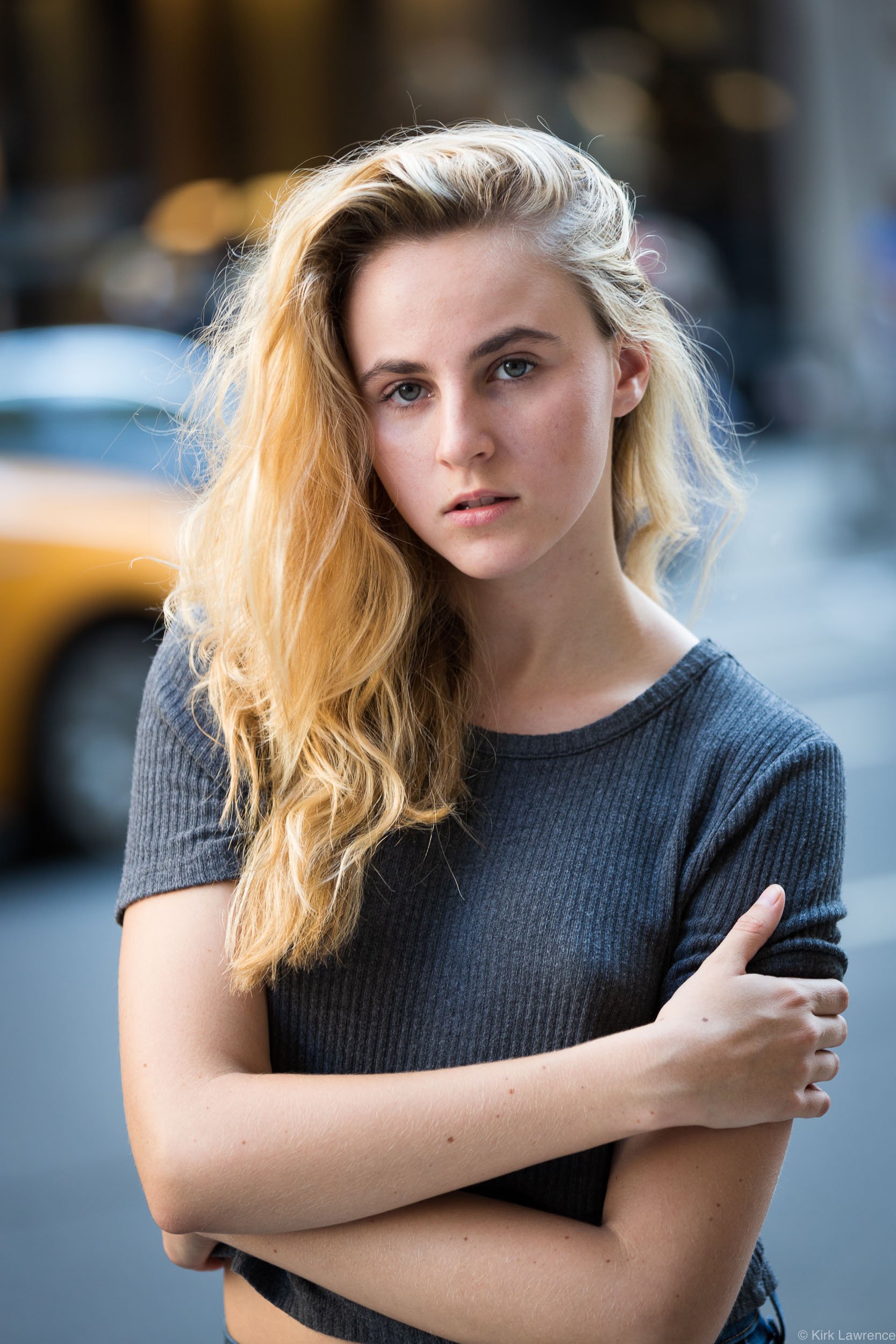 A young woman with long blonde hair and blue eyes is standing outdoors with her arms crossed, wearing a dark gray t-shirt, in front of blurred vehicles and city street background.