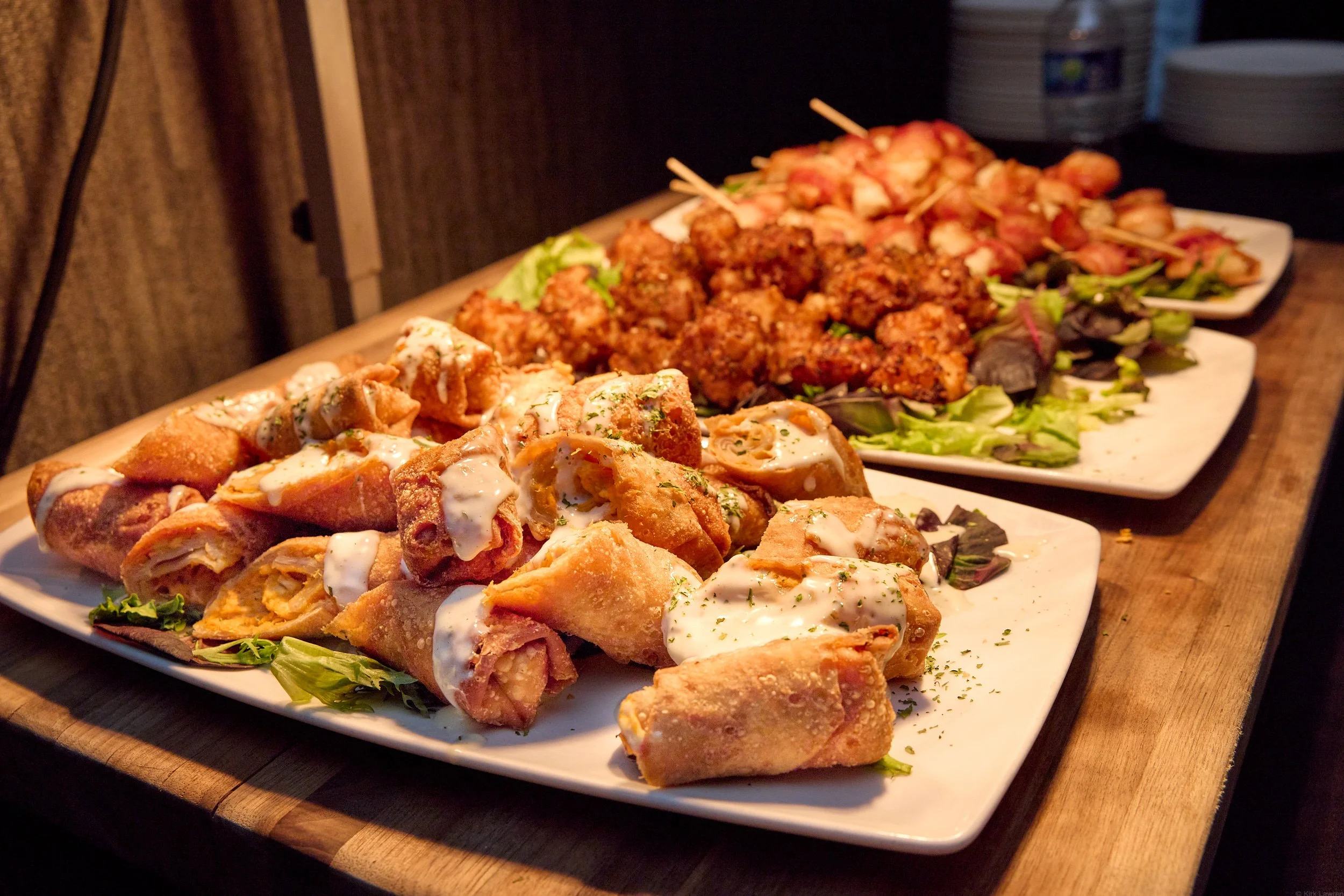 A platter of assorted appetizers including fried spring rolls topped with a white sauce, fried chicken pieces, and a variety of skewered meats and vegetables, all garnished with greens, served on a white rectangular plate.