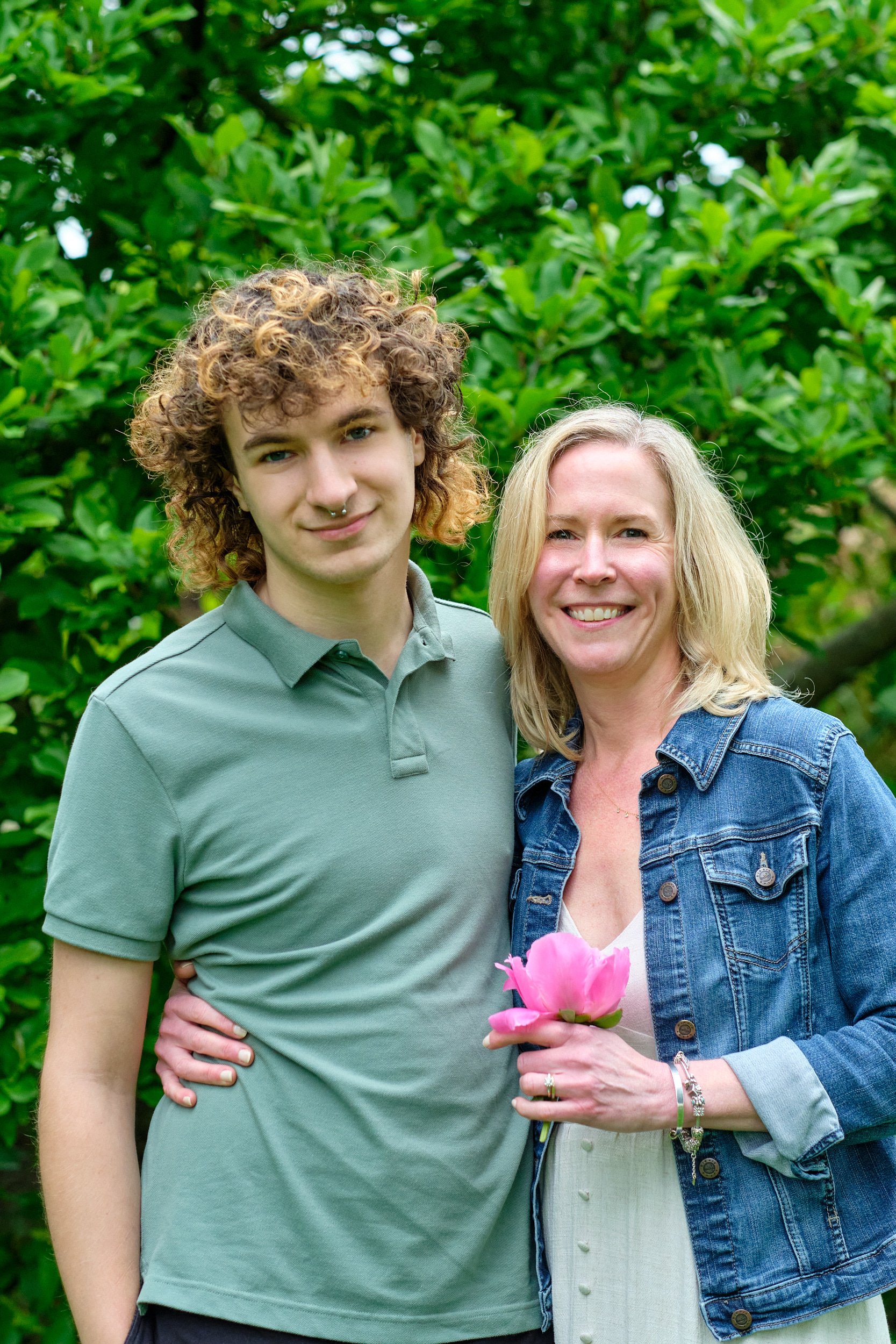 A young man with curly hair, wearing a green polo shirt, stands next to a smiling woman with blonde hair, wearing a denim jacket and holding pink flowers, in front of green foliage.