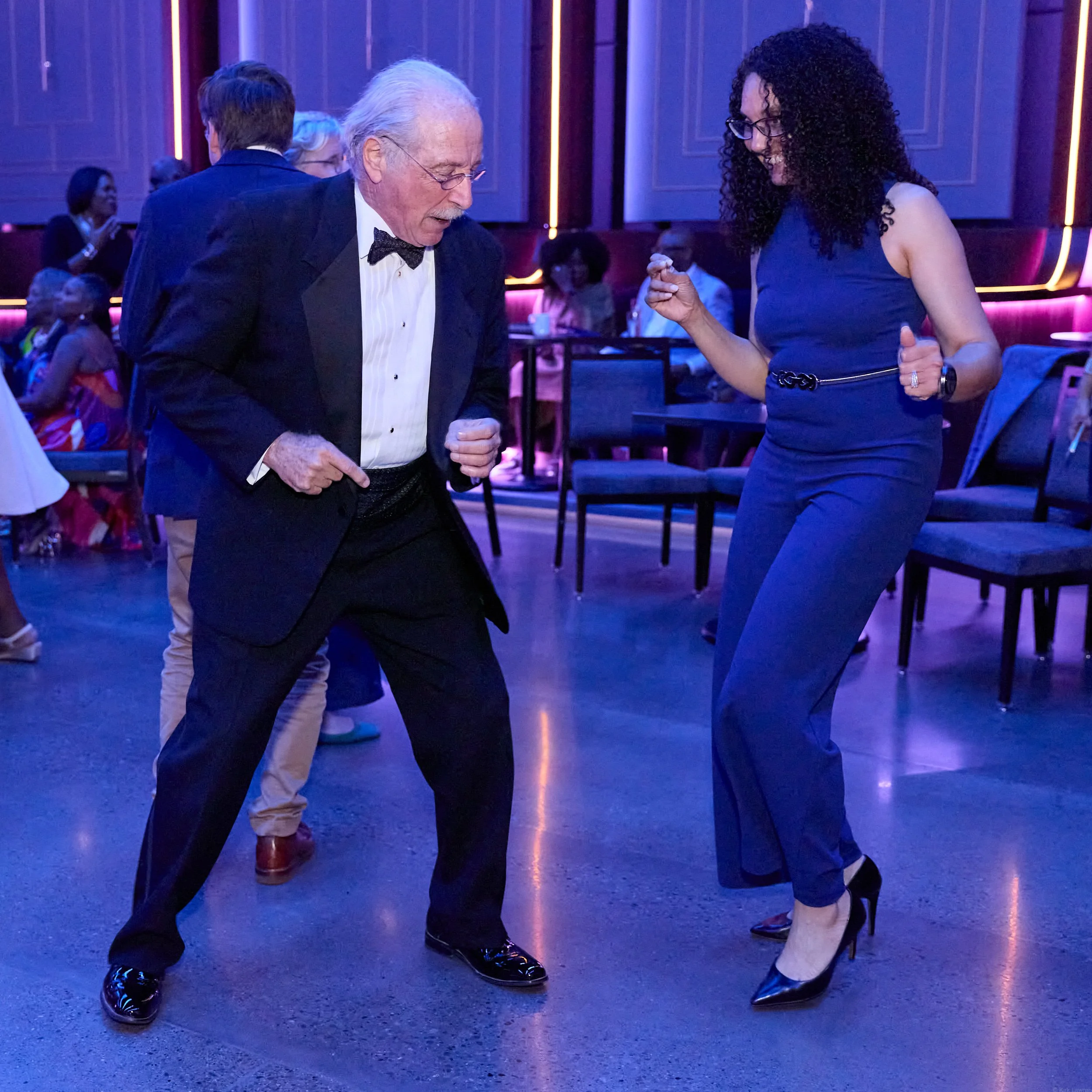 Two elegantly dressed individuals dancing together in a ballroom with purple and pink lighting, with several people watching in the background.