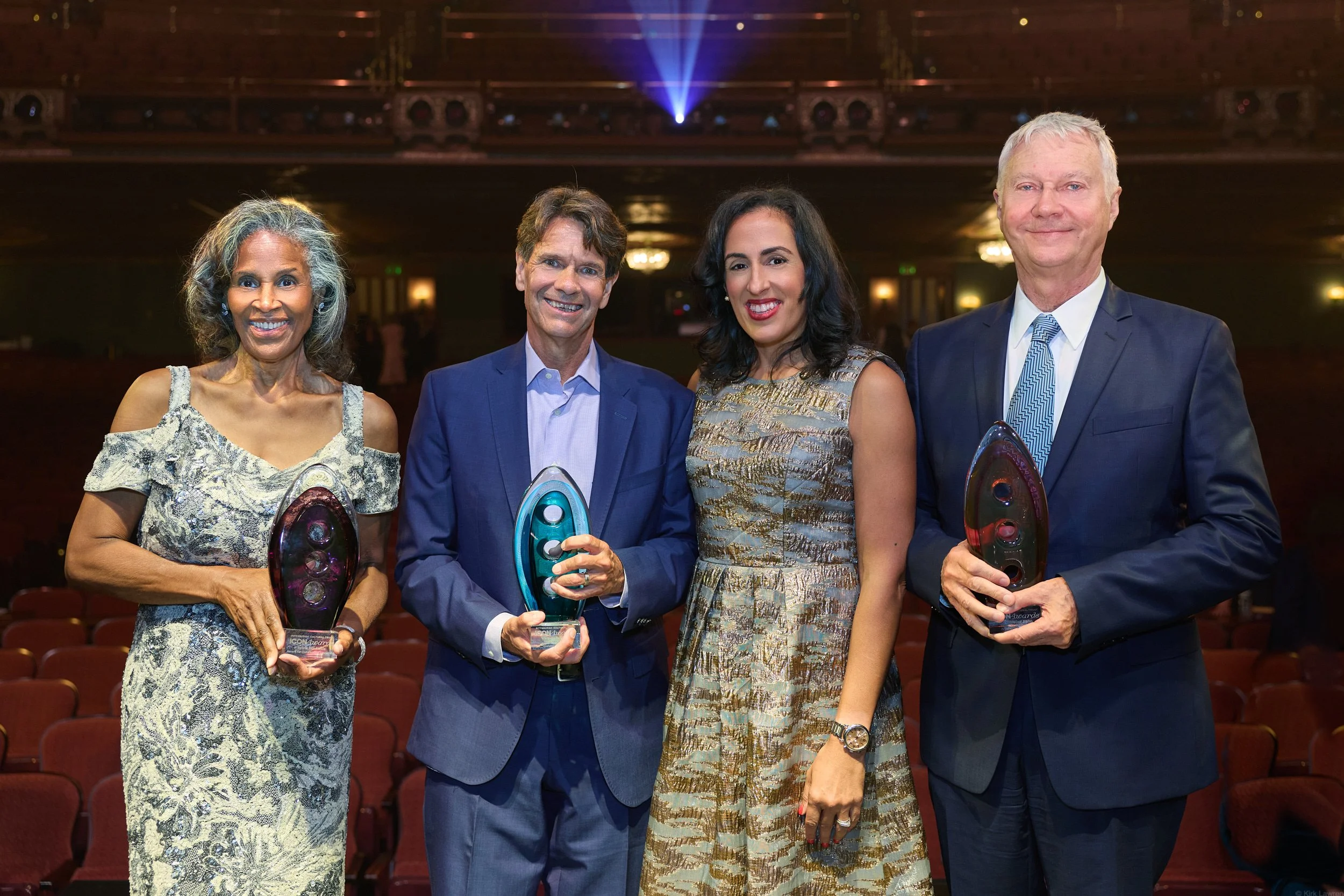 Four people, two women and two men, standing in a theater, smiling and holding awards. The woman on the left has gray curly hair and wears a lace dress. The man next to her has brown hair and wears a blue suit. The woman next to him has shoulder-leng