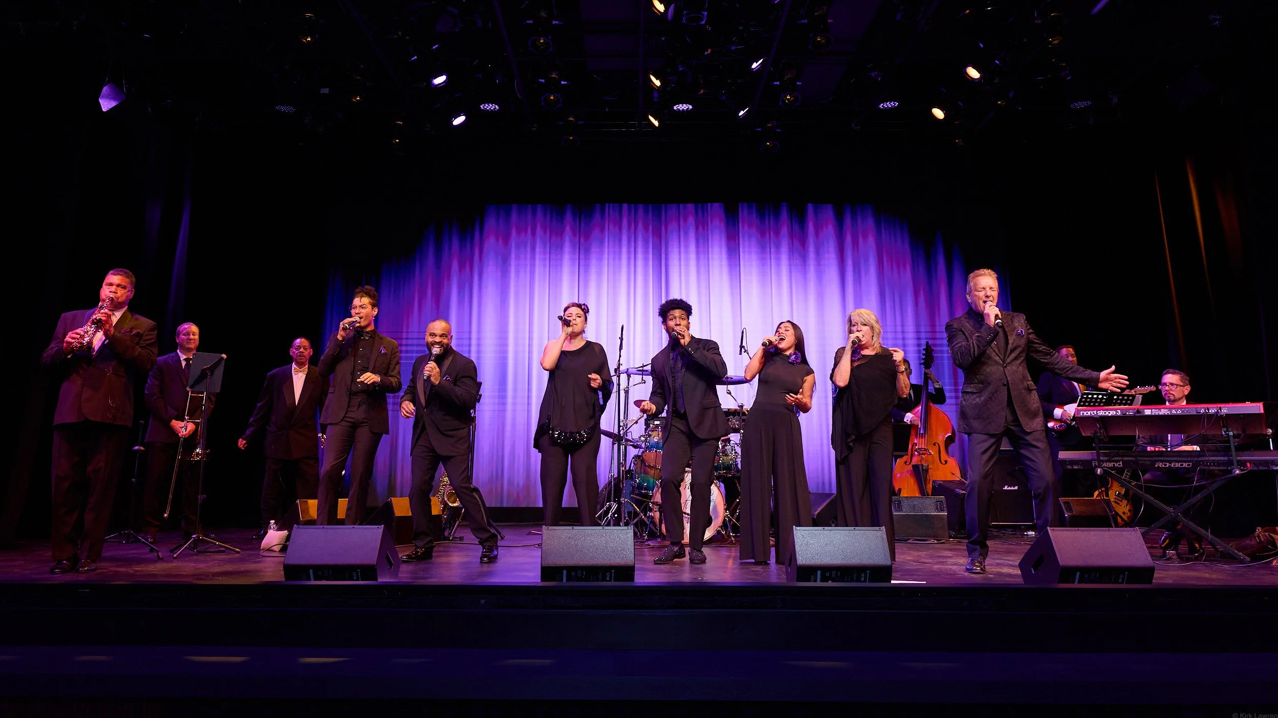 A group of performers on stage, singing and playing musical instruments during a concert with a purple curtain backdrop.