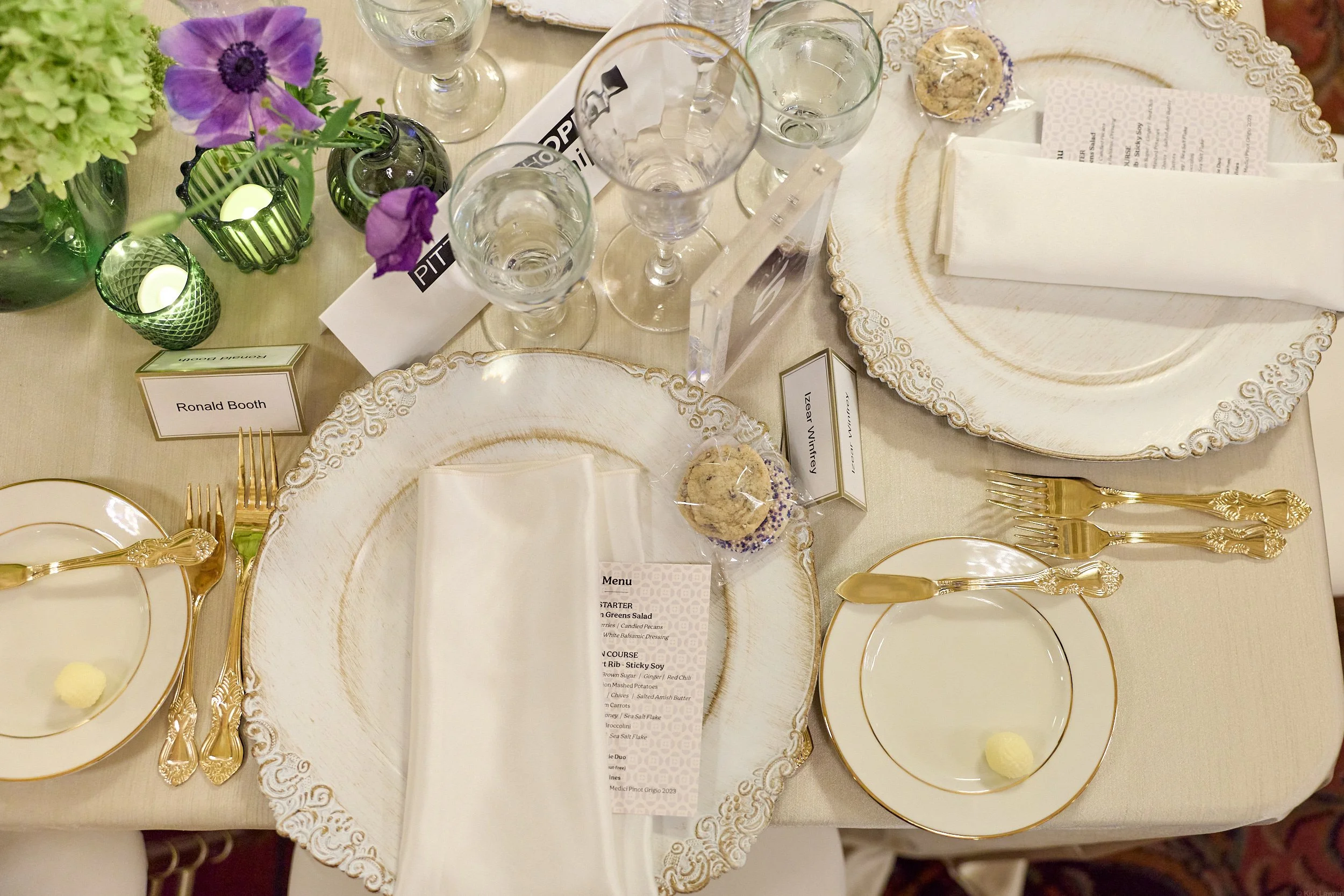 An elegant table setting featuring white and gold ornate plates, gold flatware, and champagne flutes. The table's centerpiece includes a small purple flower arrangement and green candles. A menu card with a listing of courses is placed on the table. 