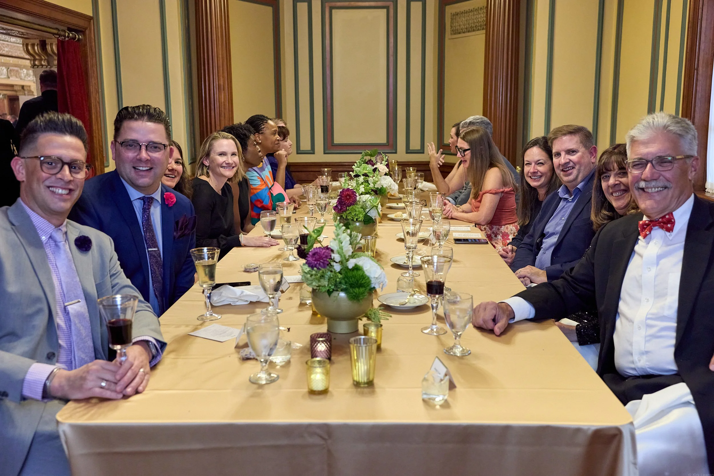 People sitting around a formal dining table, smiling, with floral centerpieces and glassware, in an elegant room with wood paneling and decorative wall features.