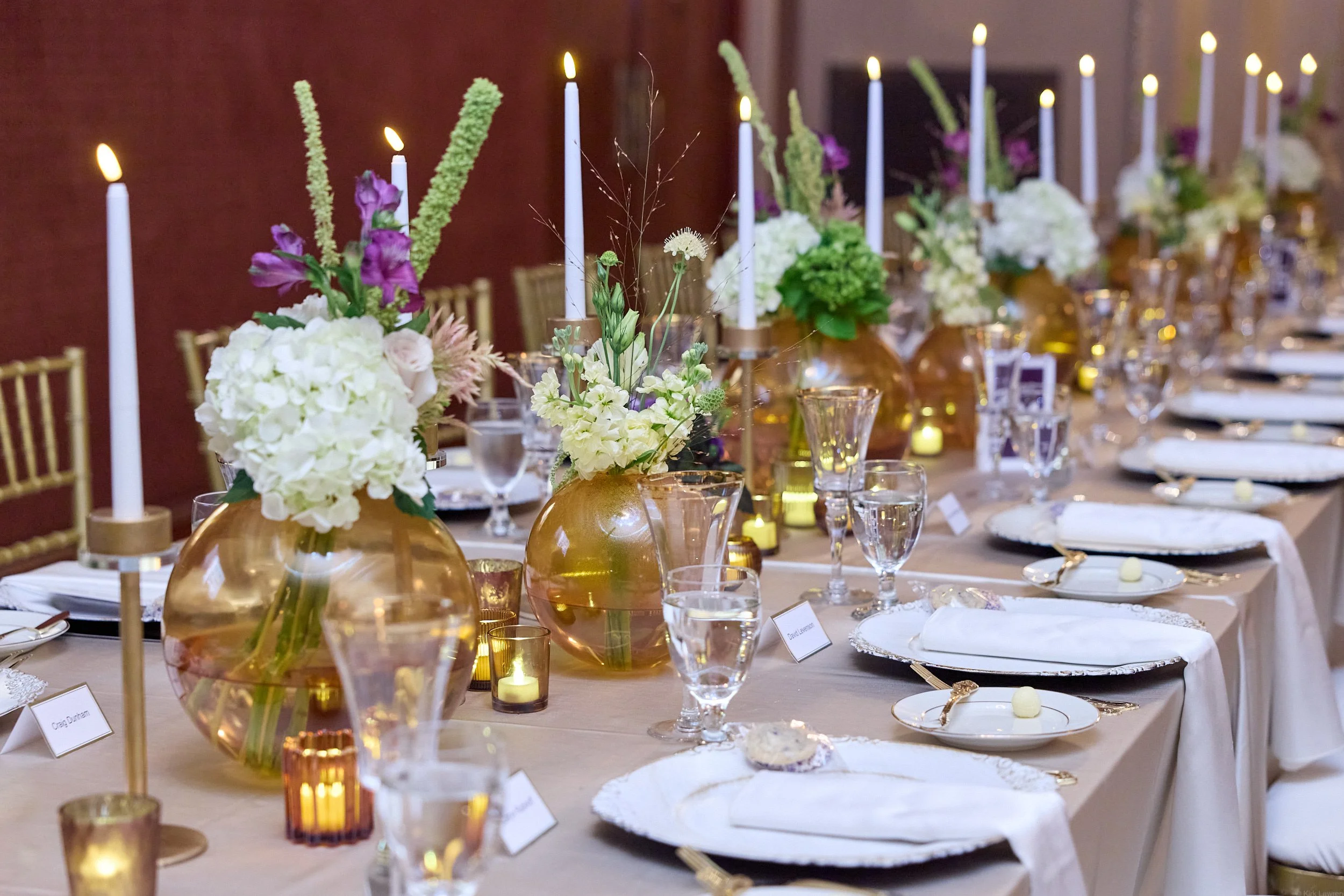 Elegant banquet table decorated with white and purple flowers, gold vases, lit candles, and gold accents, set for a formal event.