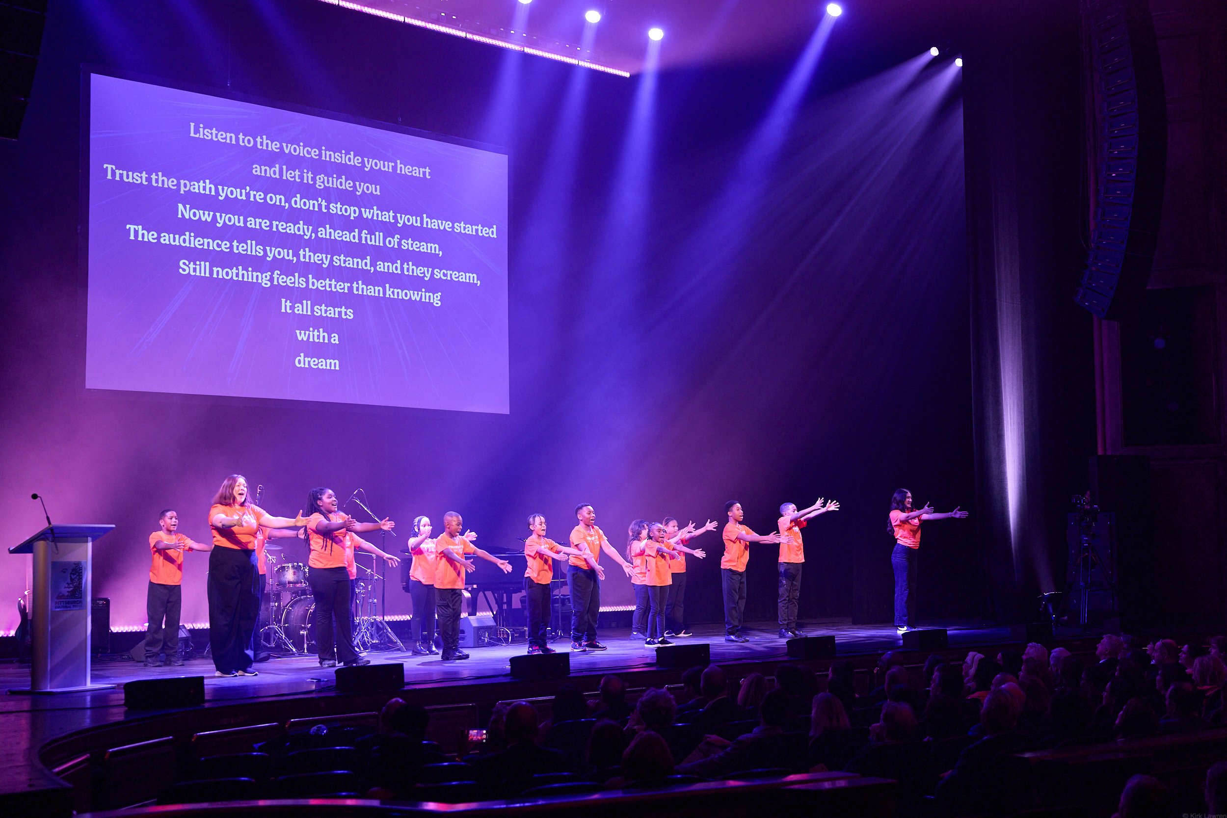 Children performing on stage with a large screen displaying lyrics in the background, purple and blue stage lighting