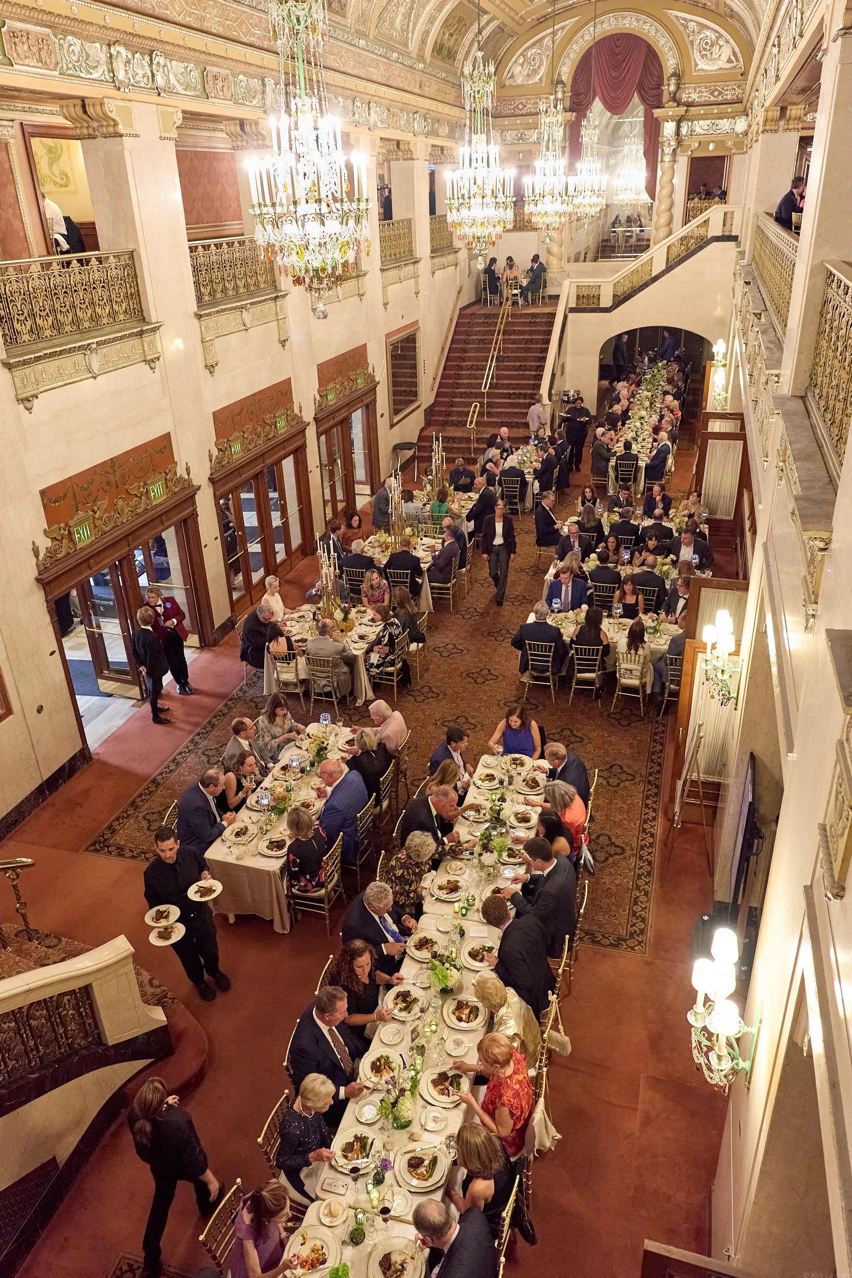 A grand ballroom with ornate gold and white decor, large chandeliers, and a long banquet table filled with guests enjoying a formal dinner.