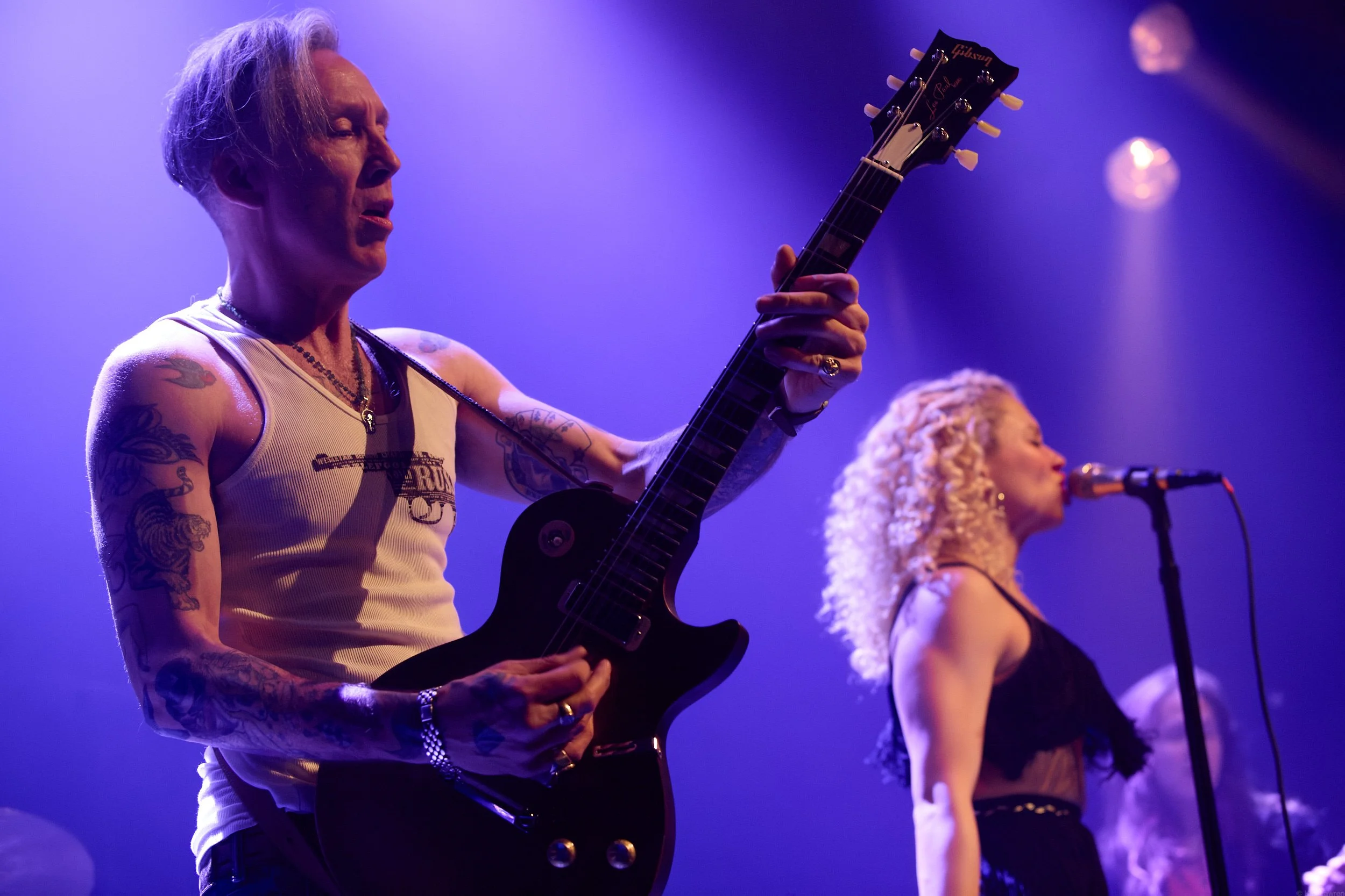 A male musician with tattoos on his arms, wearing a white tank top, playing an electric guitar on stage with purple lighting. A female singer with blonde, curly hair is standing to his right, singing into a microphone.