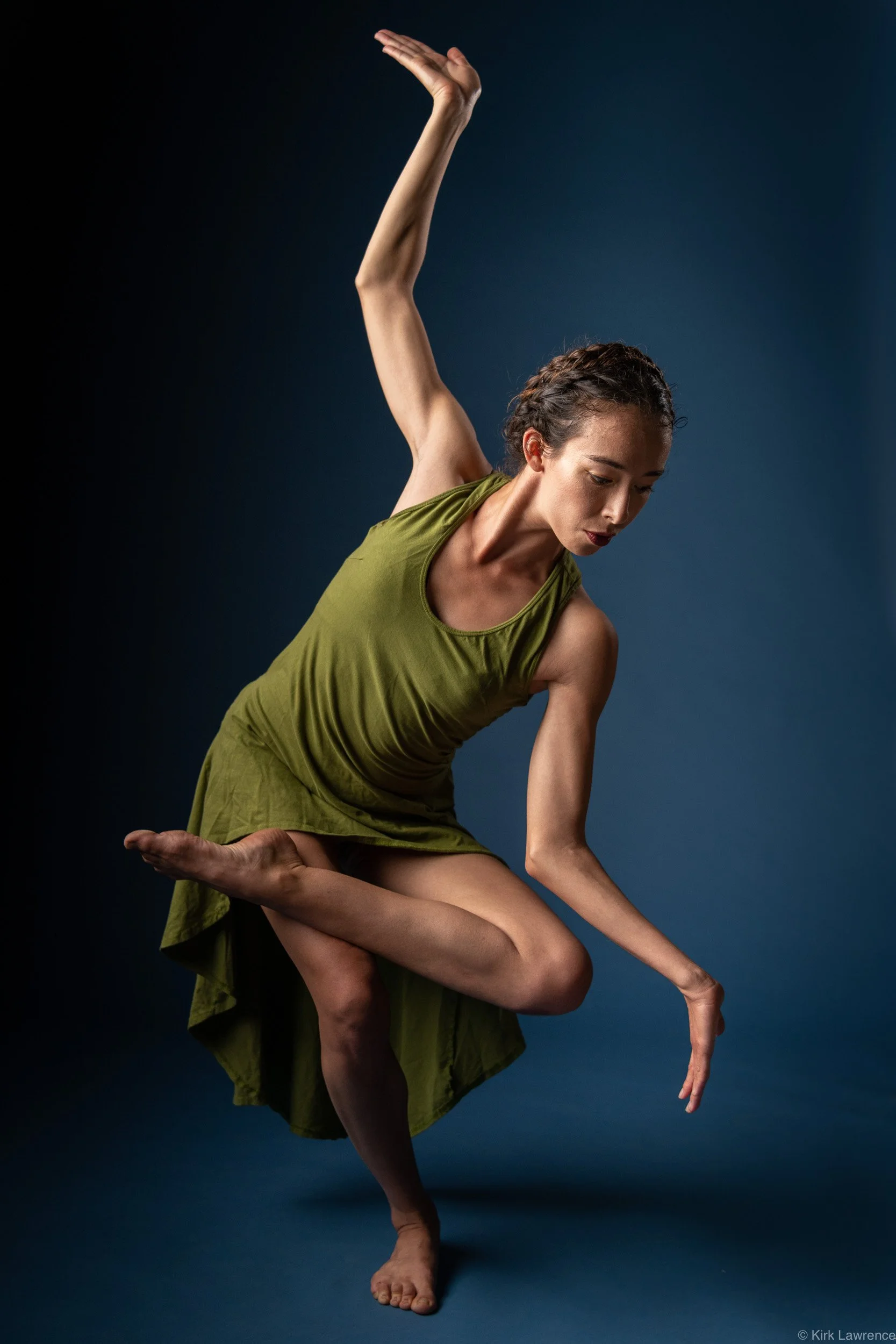 A woman performing a ballet pose, balancing on one leg with her other leg bent and arm extended, against a dark blue background.