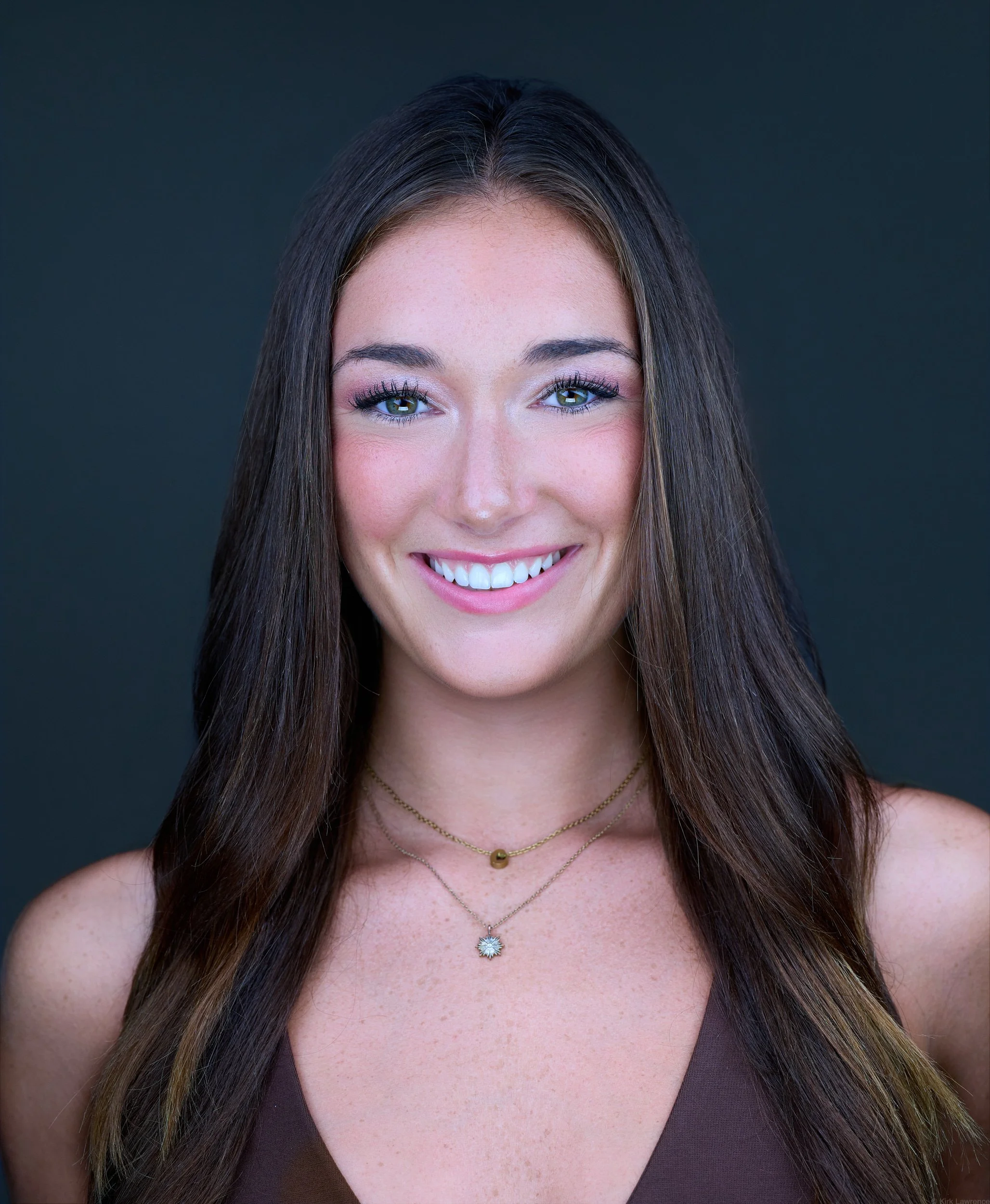 Close-up of a young woman with long brown hair, smiling, wearing makeup, layered necklaces, and a brown top, against a dark background.